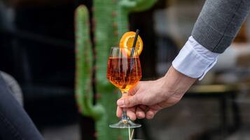 A hand holding a cocktail glass with an orange slice, a glass of olives, and a reflection on a glass table.