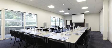 Un salón de conferencias con una larga mesa, sillas, botellas de agua, tarjetas de identificación y una pantalla para proyector.