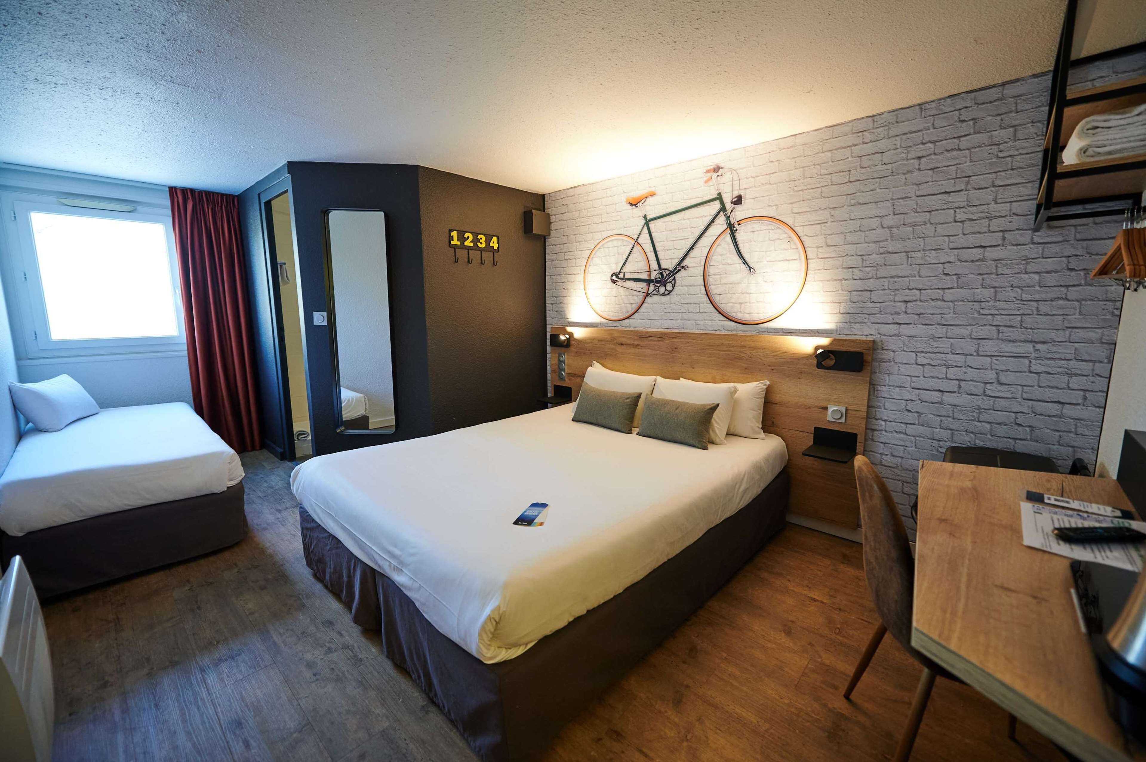 A hotel room with a large bed, two smaller beds, a wooden desk, and a bicycle mounted on the wall.