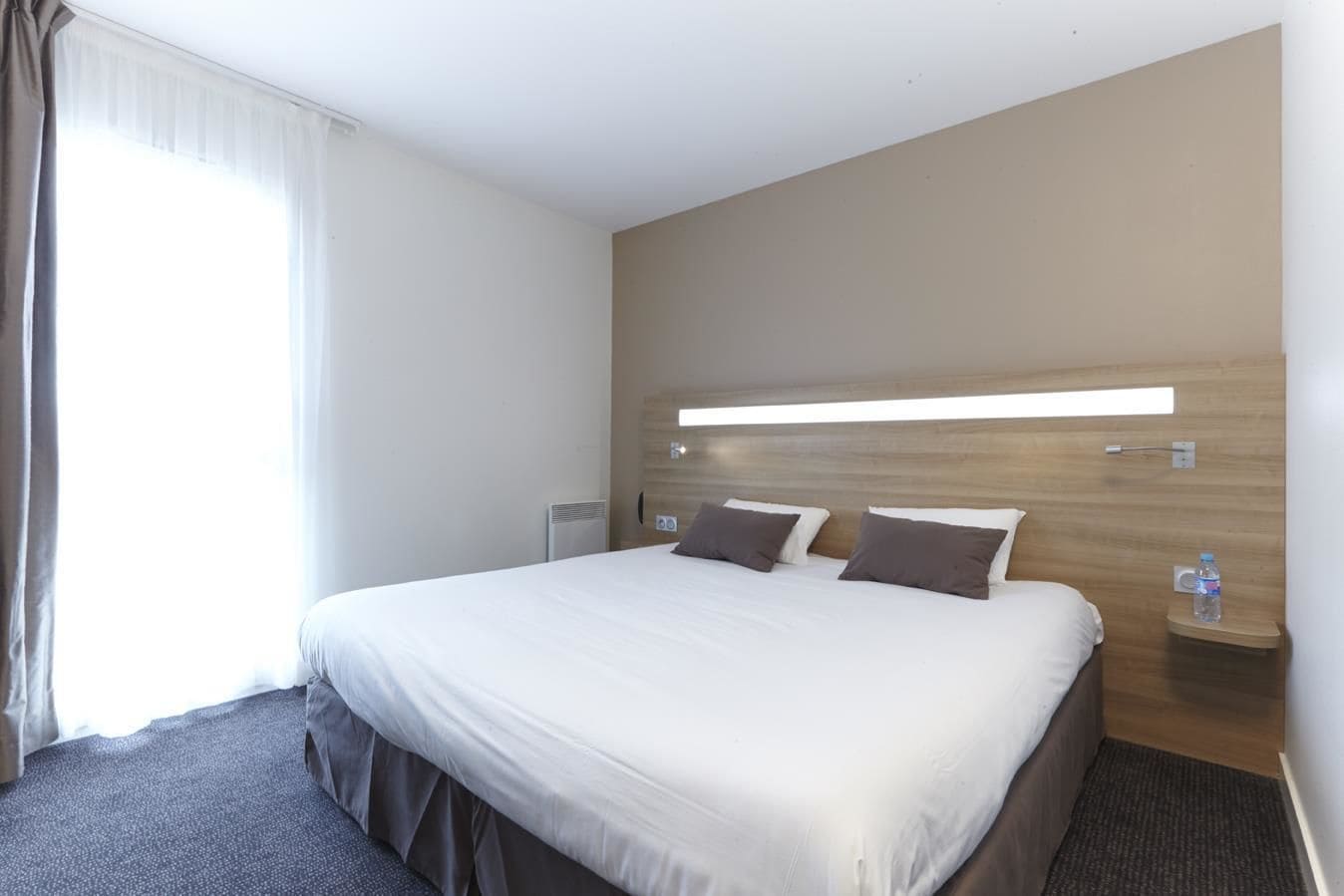 Habitación de hotel con una cama grande, una mesita de noche con una botella de agua y una lámpara fijada a la pared.