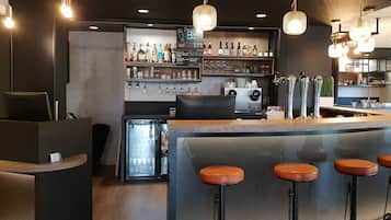Una zona de bar con mostrador, taburetes, un refrigerador y estantes con botellas y vasos.