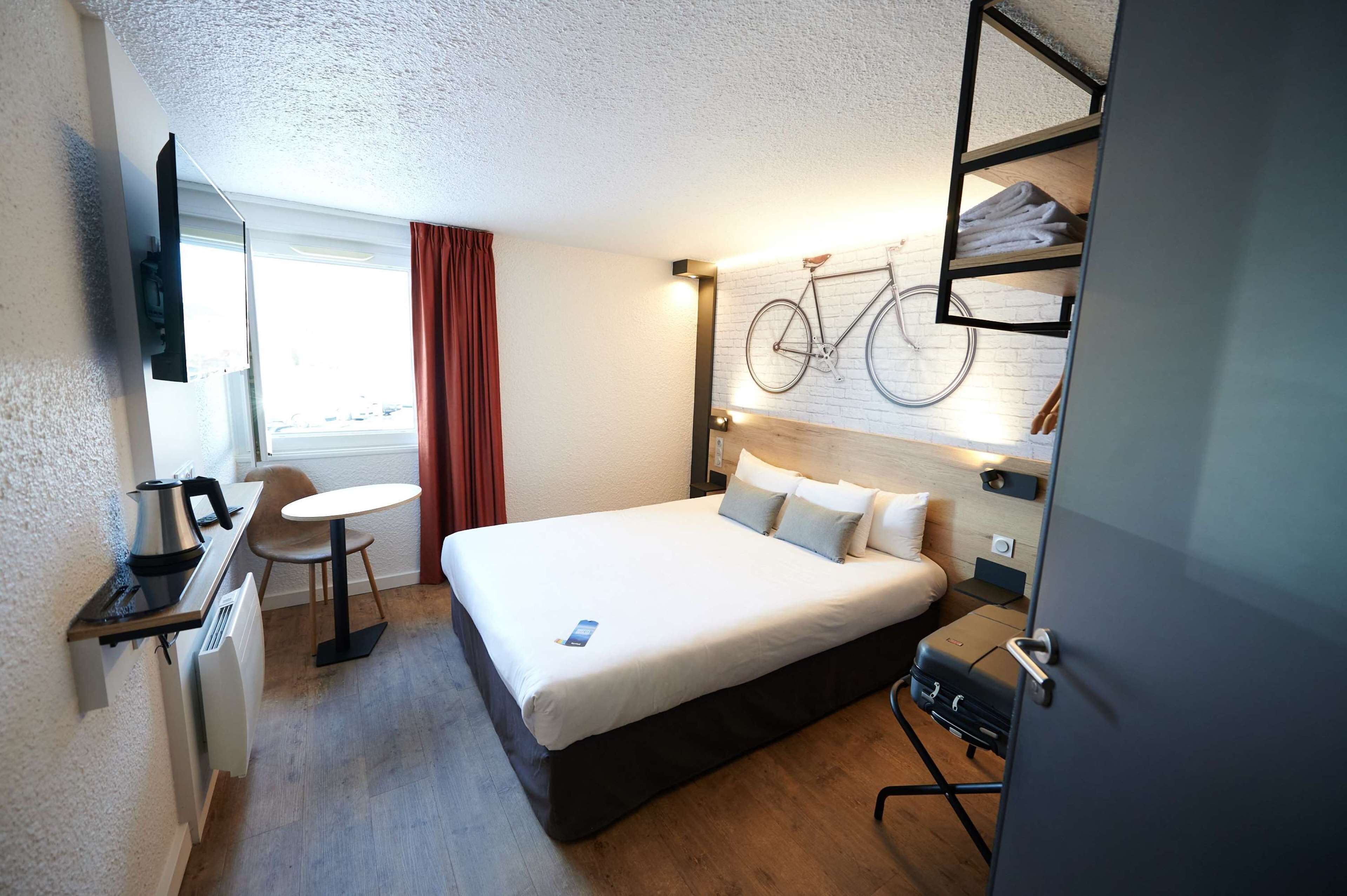 Une chambre d’hôtel comprenant un lit, un bureau avec une bouilloire, une chaise, une petite table, un vélo fixé au mur et un radiateur.