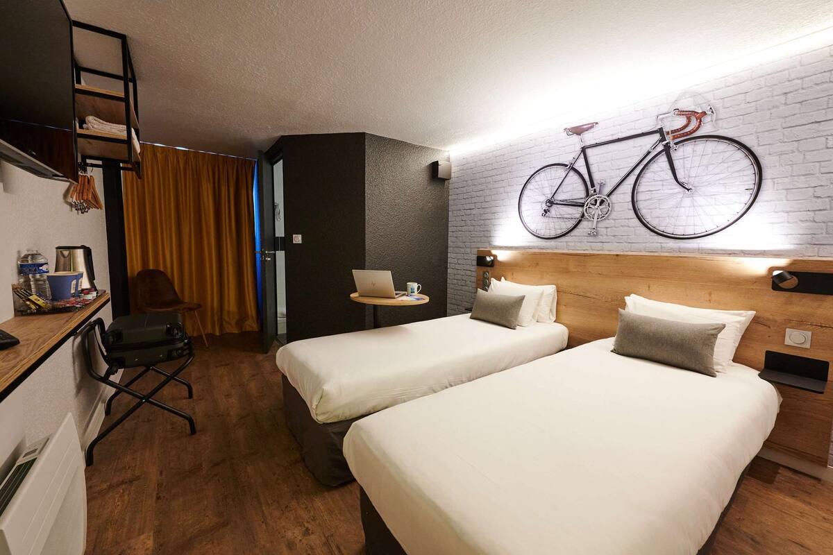 Habitación de hotel con dos camas, un escritorio con una computadora portátil, una bicicleta colgada en la pared y un cabecero de madera.