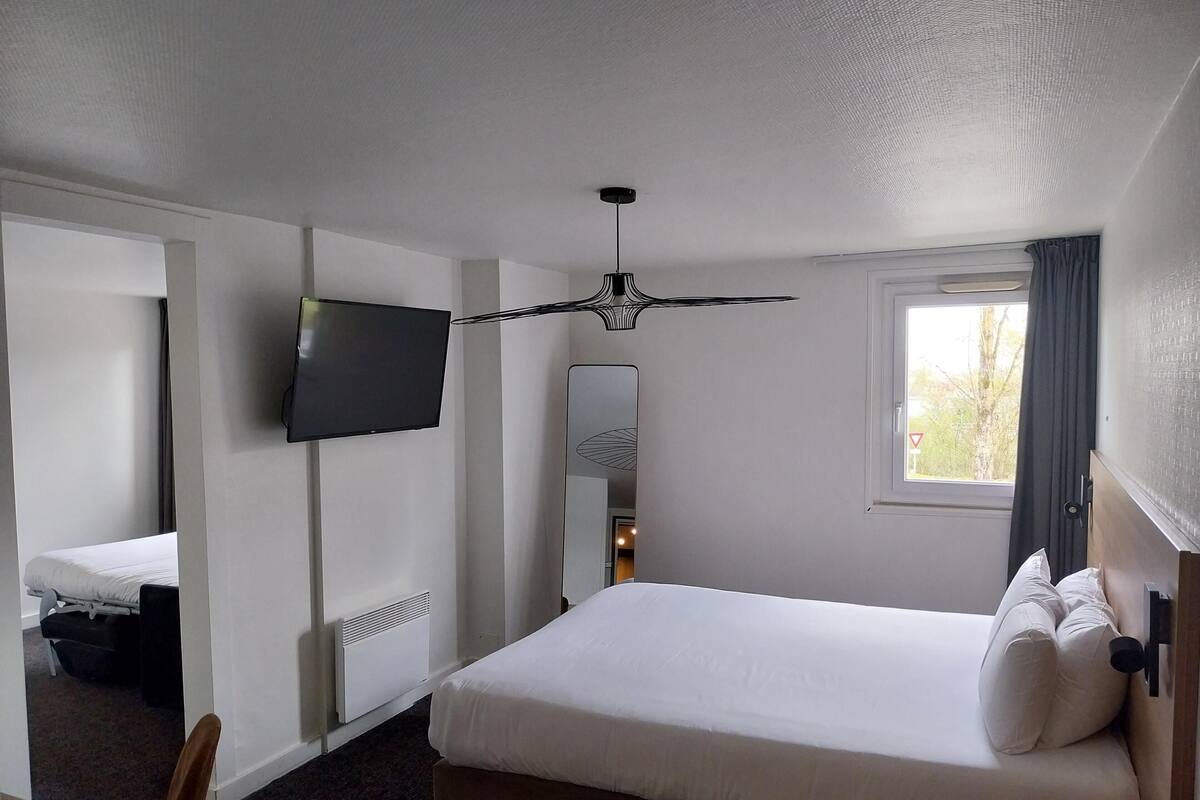 Habitación de hotel con cama, televisión, ventilador de techo, ventana con cortinas y radiador.