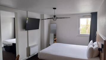 A hotel room with a bed, a TV, a ceiling fan, a window with curtains, and a radiator.