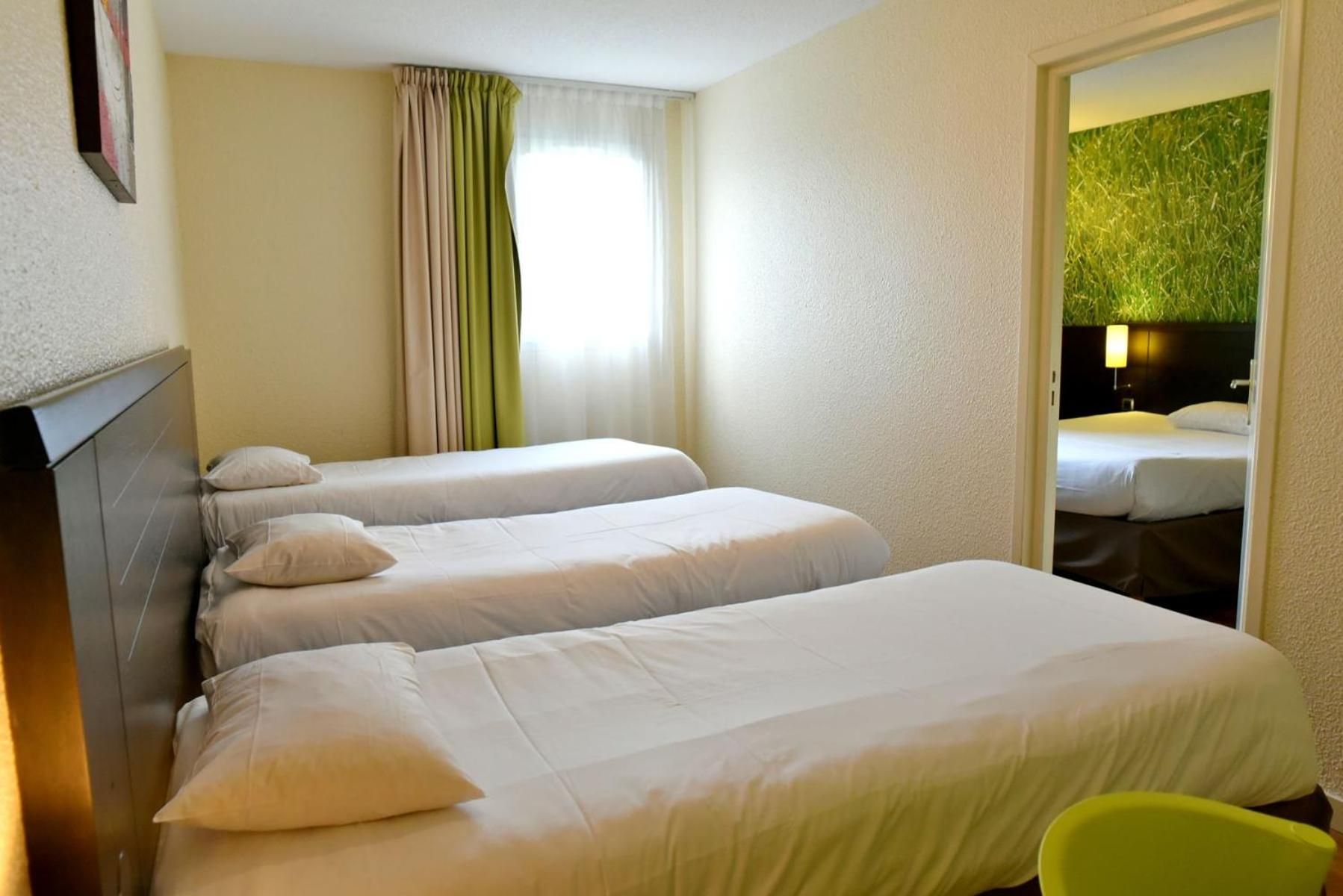 Une chambre d’hôtel avec deux lits, un fauteuil vert et une vue sur un mur verdoyant, visible à travers une porte ouverte.