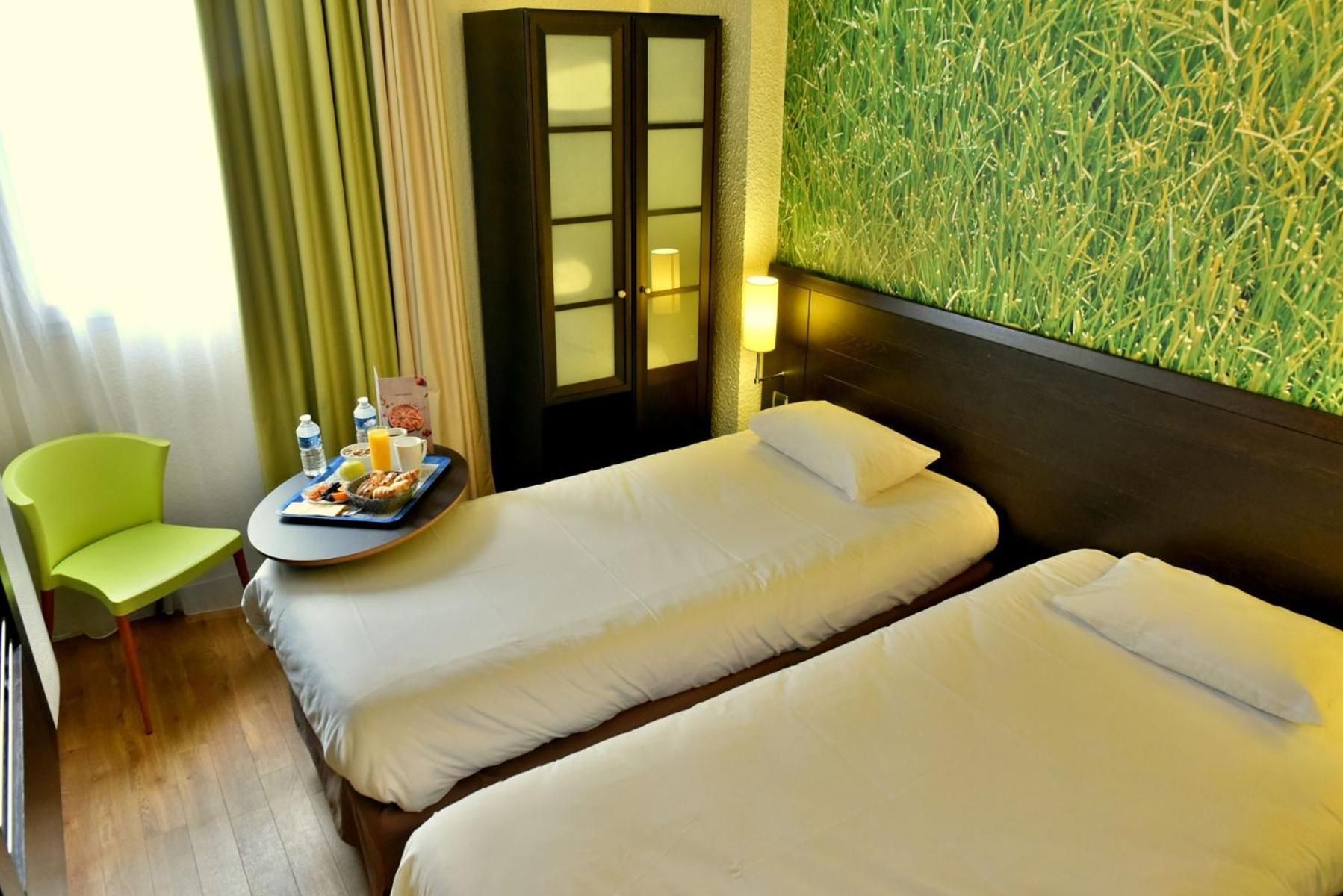 Habitación de hotel con dos camas, una mesita con desayuno, una silla verde y una pared con diseño que imita hierba.