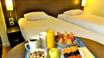 Una habitación de hotel con dos camas, una bandeja de desayuno con croissants, frutas, un vaso de zumo de naranja, un bol de cereales y una taza de café.
