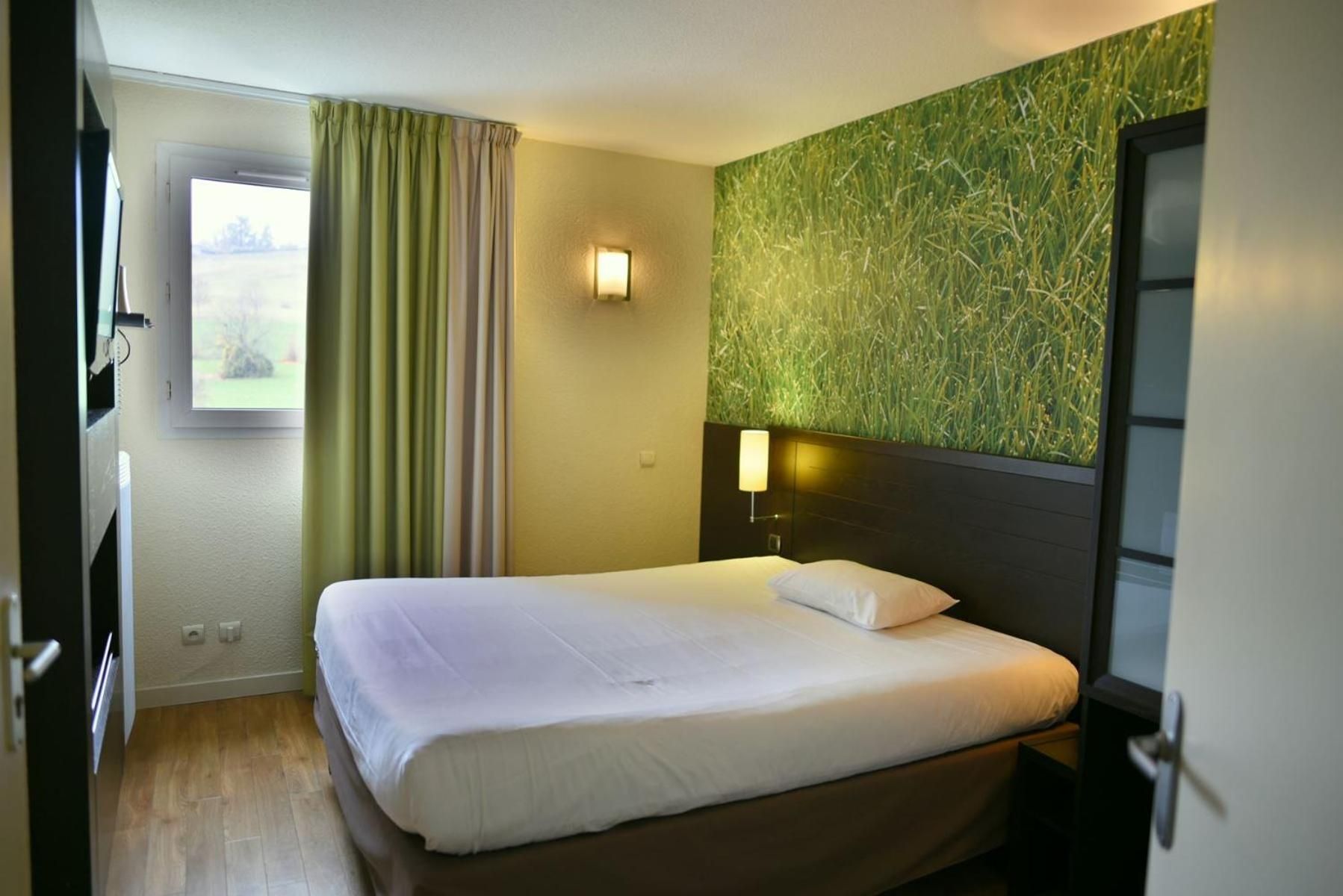 A hotel room with a bed, a window with curtains, a wall-mounted lamp, and a door.
