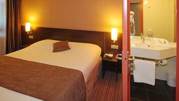Quarto de hotel com uma cama grande, luminárias de cabeceira, mesa de cabeceira, banheiro com pia, espelho e porta-toalhas.