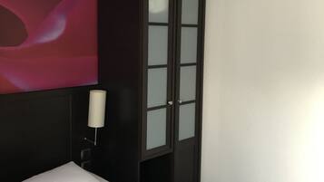 A hotel room with a bed, a dark wooden wardrobe with glass doors, a white wall, and a partial view of a pink abstract mural.