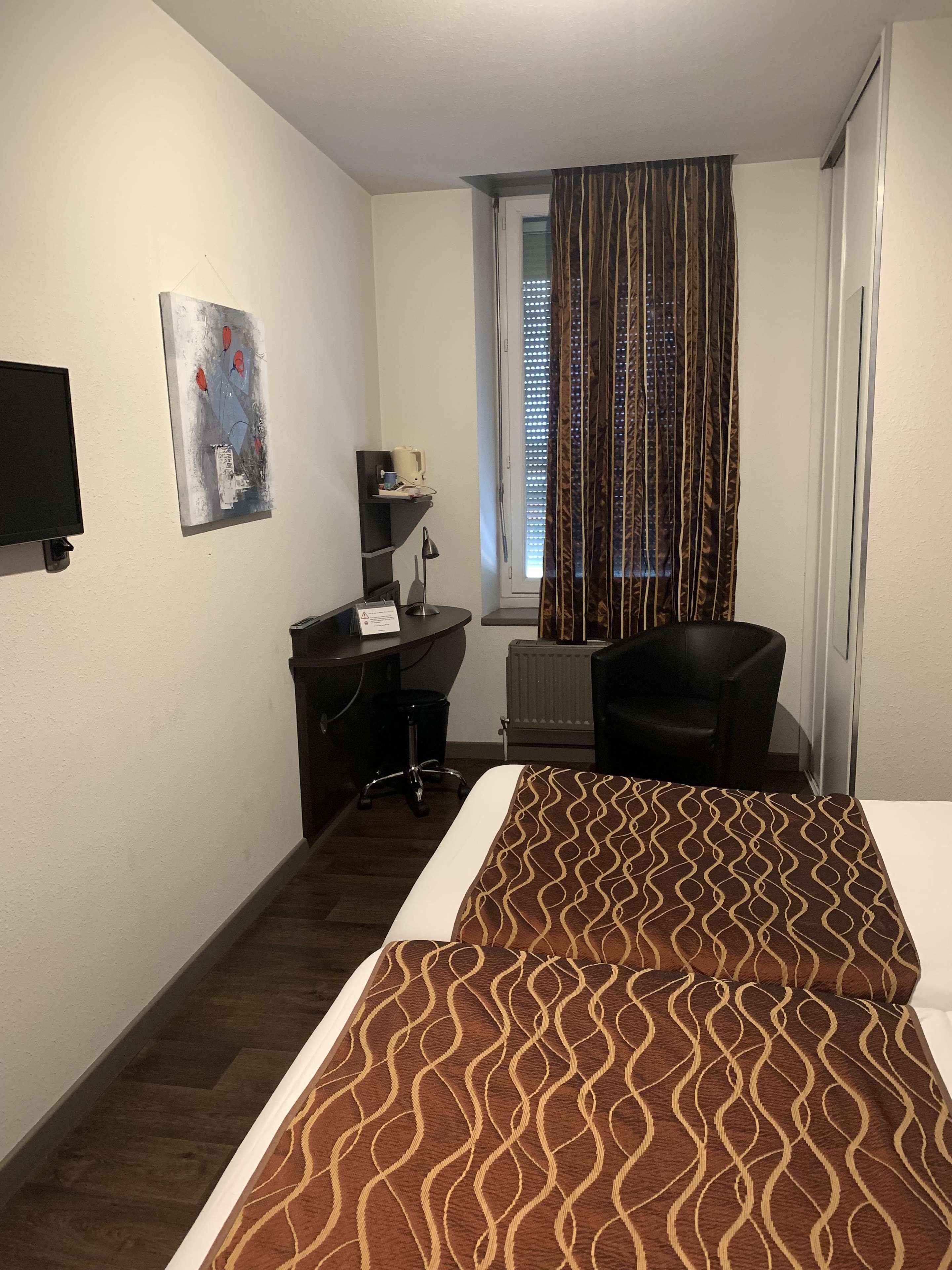 A hotel room with a bed, a desk, a chair, a TV, a window with curtains, and a radiator.