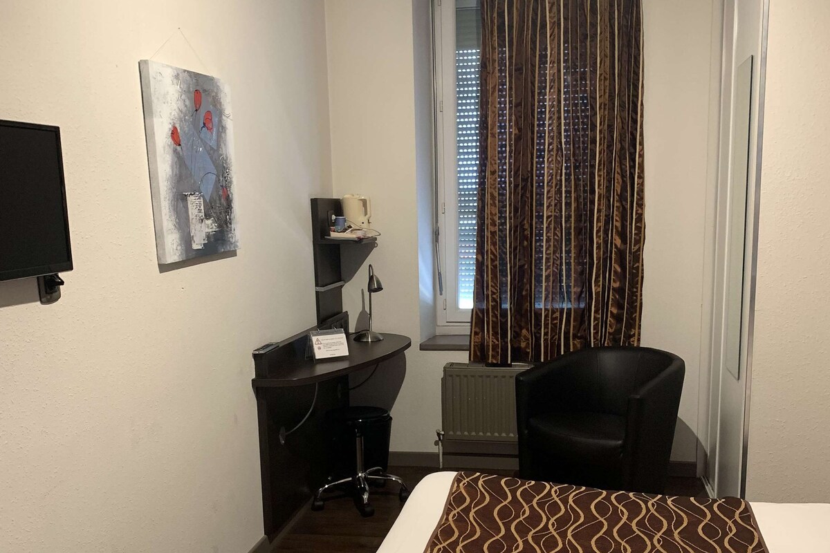 A hotel room with a bed, a desk, a chair, a TV, a window with curtains, and a radiator.
