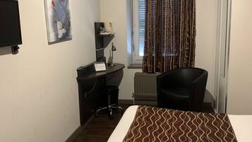 A hotel room with a bed, a desk, a chair, a TV, a window with curtains, and a radiator.