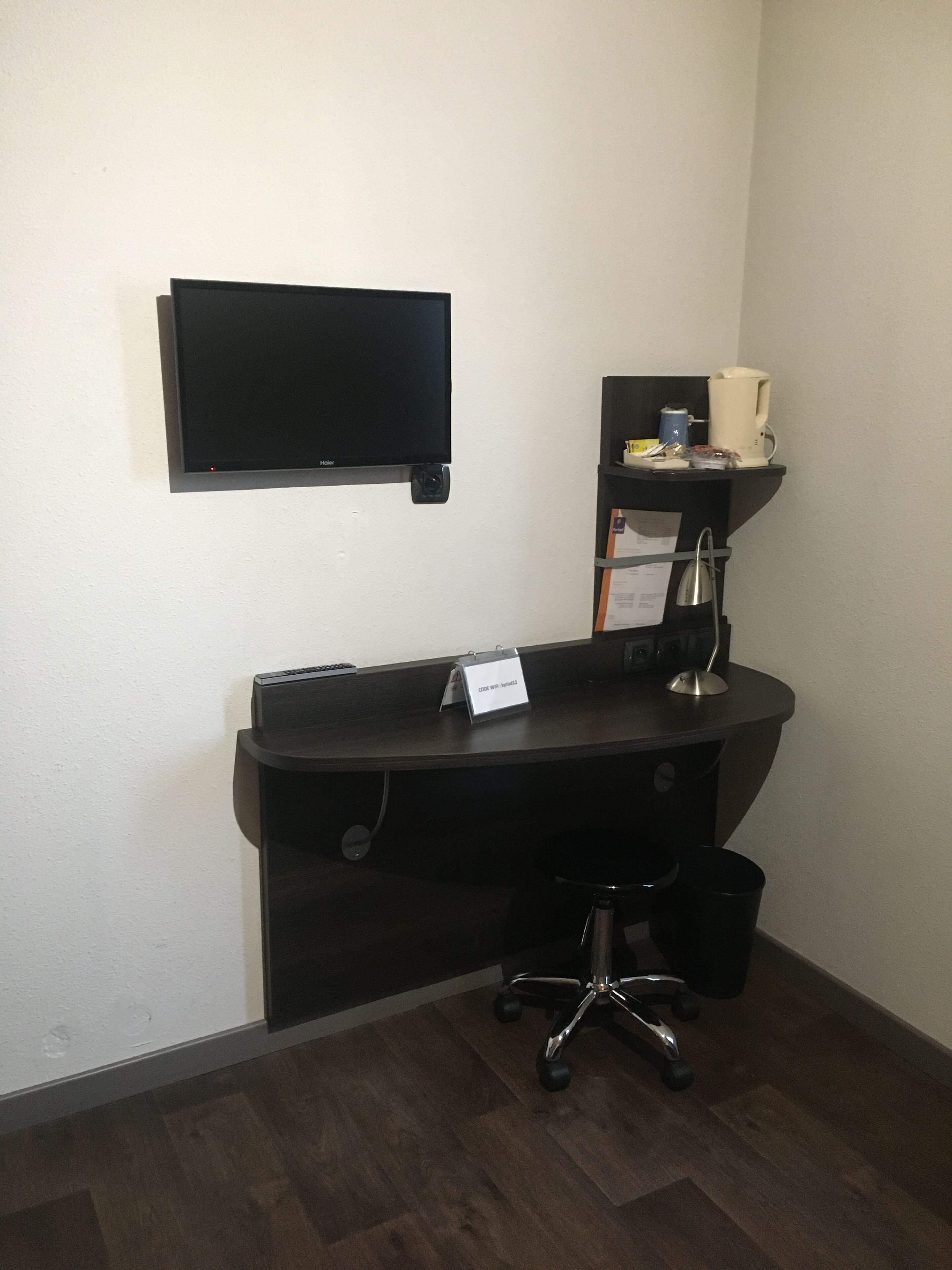 Un pequeño escritorio de madera oscura con un televisor de pantalla plana montado encima, una cafetera y un estante pequeño con una taza y otros objetos.