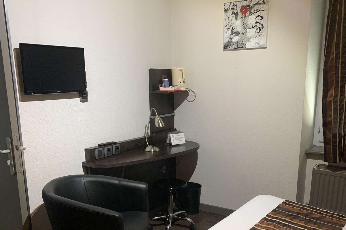 A hotel room with a bed, a desk, a chair, a television, and a painting on the wall.