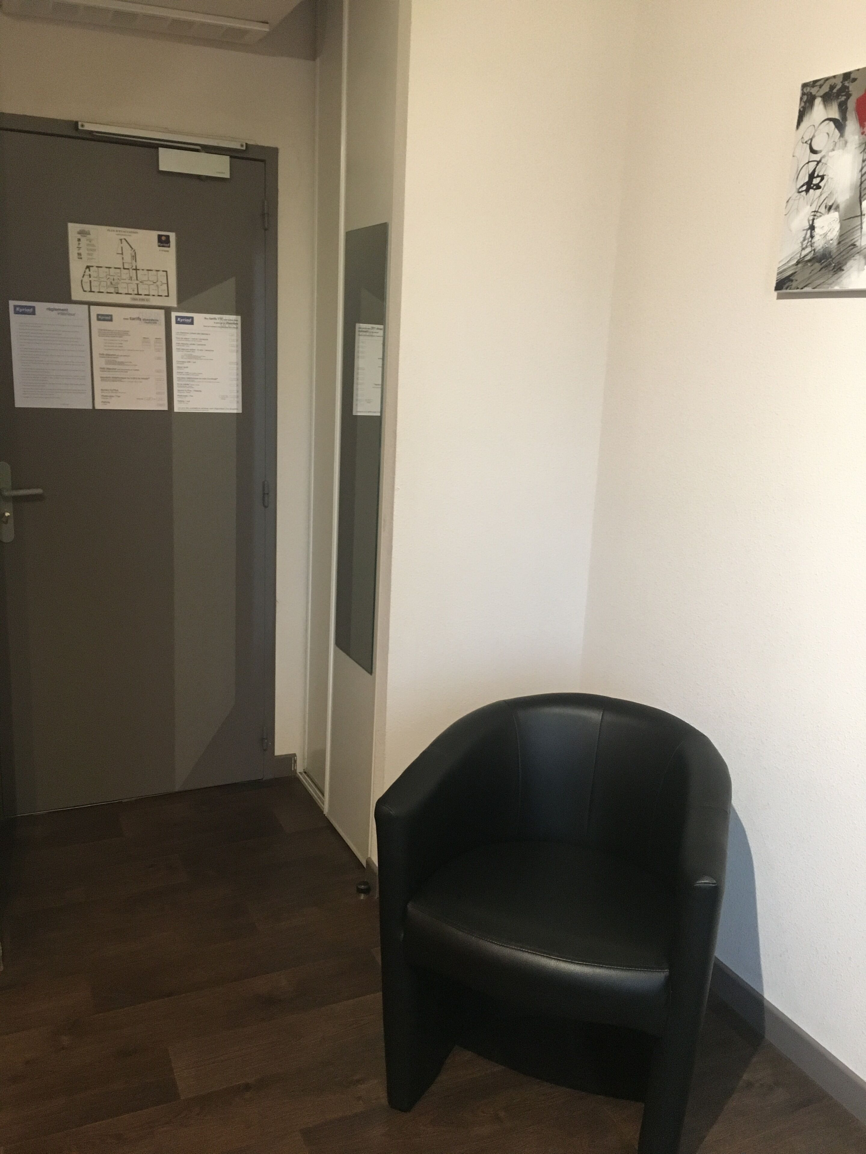 A room with a black chair, a door with notices, and a wall with a framed picture.
