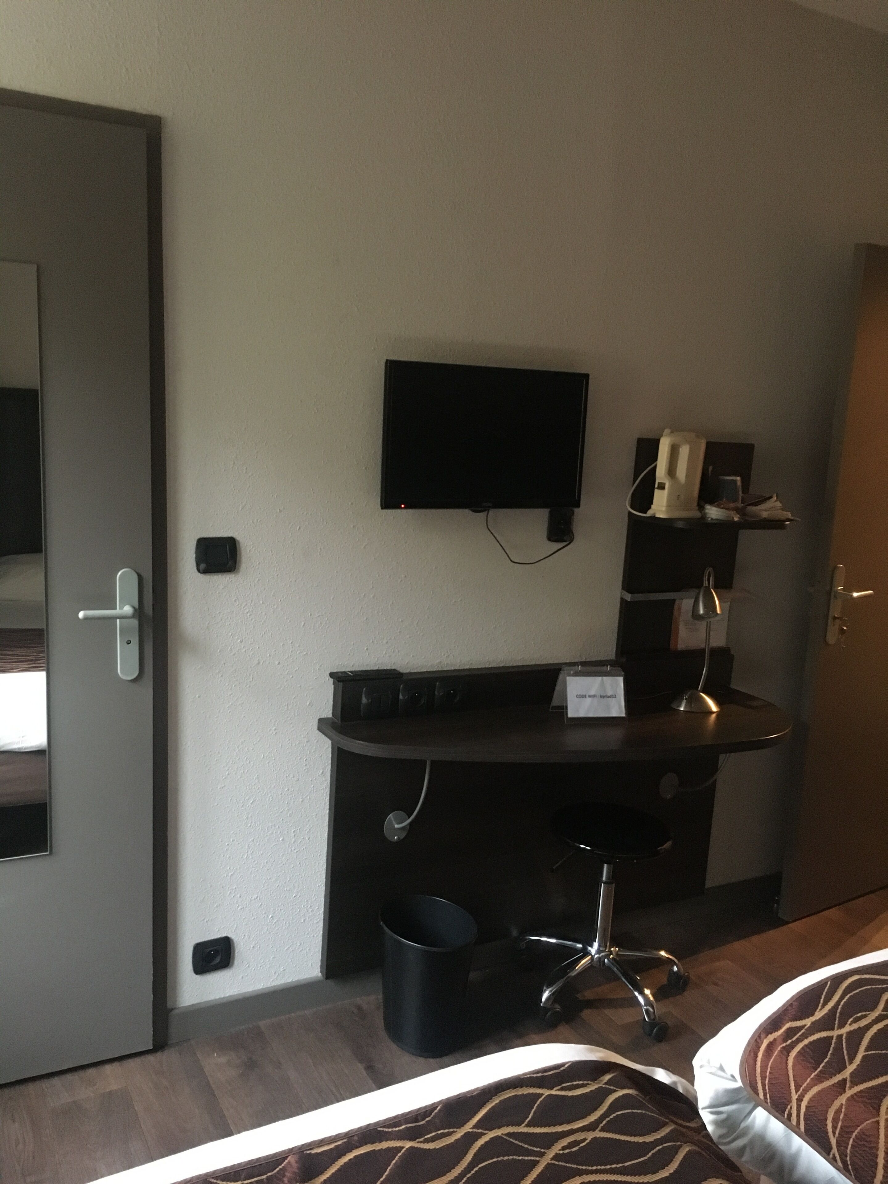 Een hotelkamer met een bed, een bureau met een computer, een aan de muur bevestigde televisie en een kleine prullenbak.