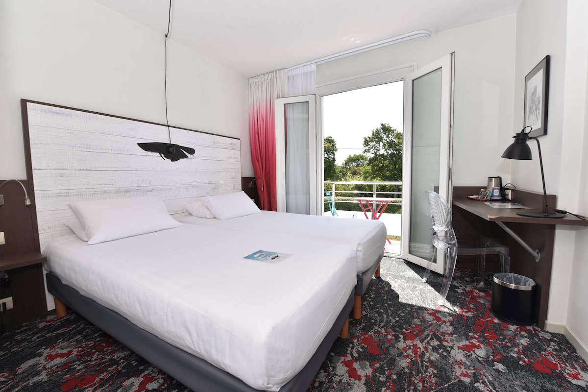 A hotel room with a large bed, a desk with a chair, a small trash bin, and a balcony with a chair.