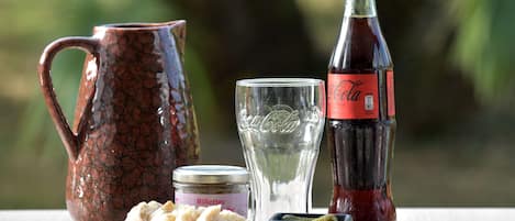 Un entorno rústico al aire libre con una jarra de cerámica, una botella de Coca-Cola, un vaso y un frasco de galletas sobre una mesa de madera.