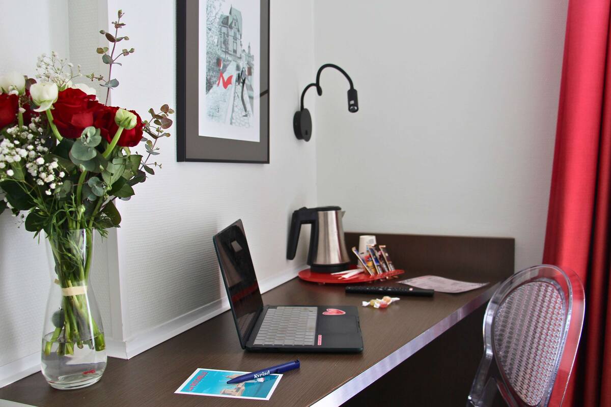 Un escritorio con una laptop, un florero, un cuadro enmarco, una cafetera, audífonos y una silla.