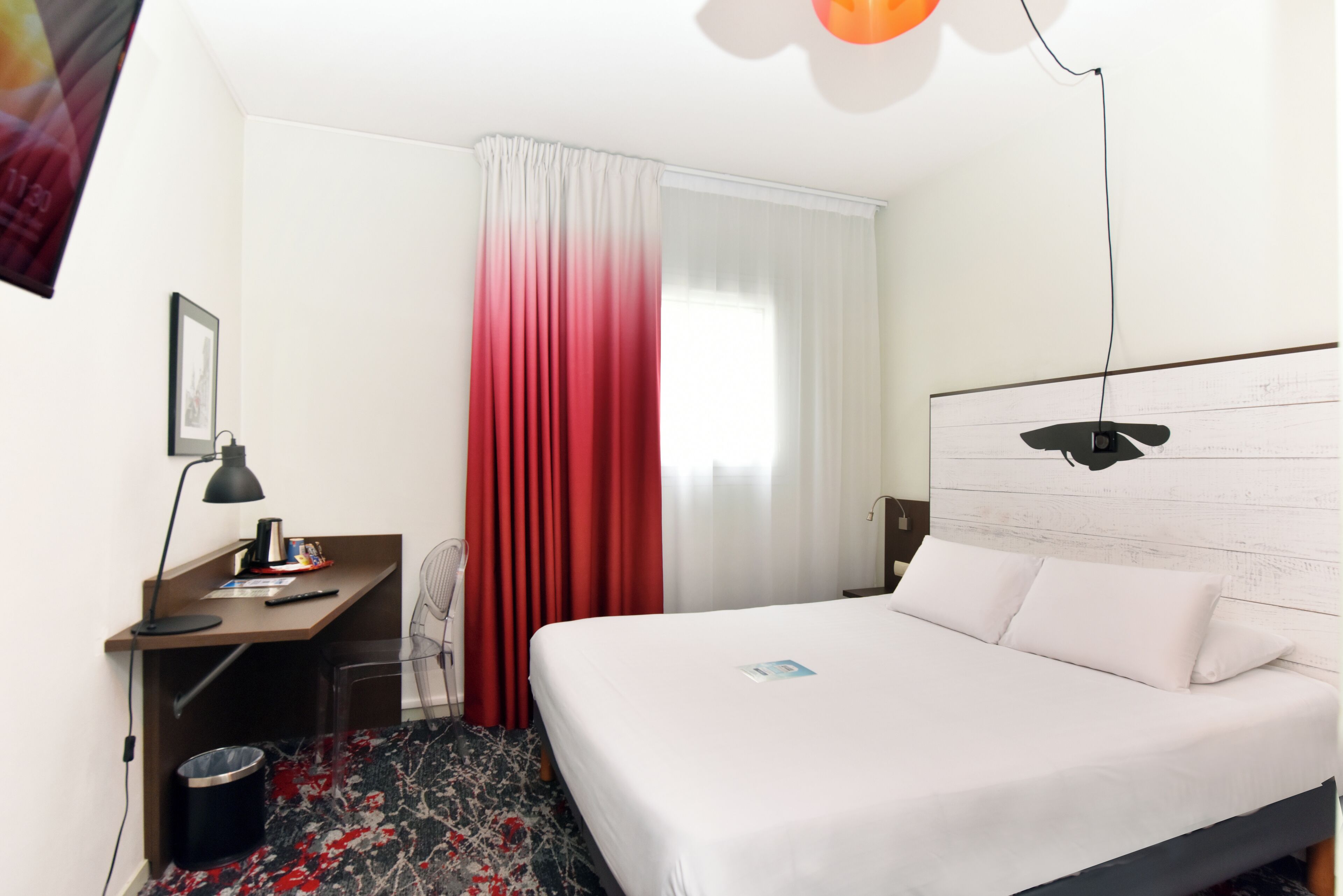 A hotel room with a bed, a desk, a chair, a lamp, and a window with curtains.