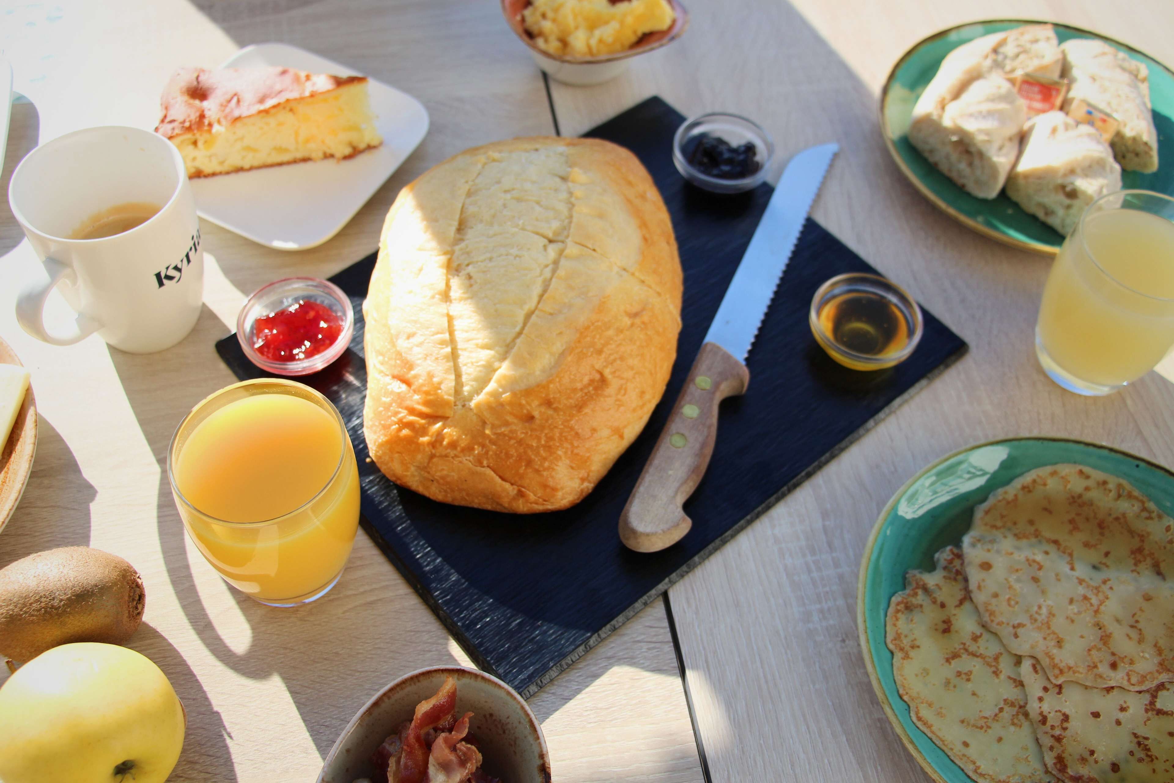 Une table de petit-déjeuner avec une miche de pain, une assiette de crêpes, une tasse de café, un verre de jus d’orange et un bol de fruits.