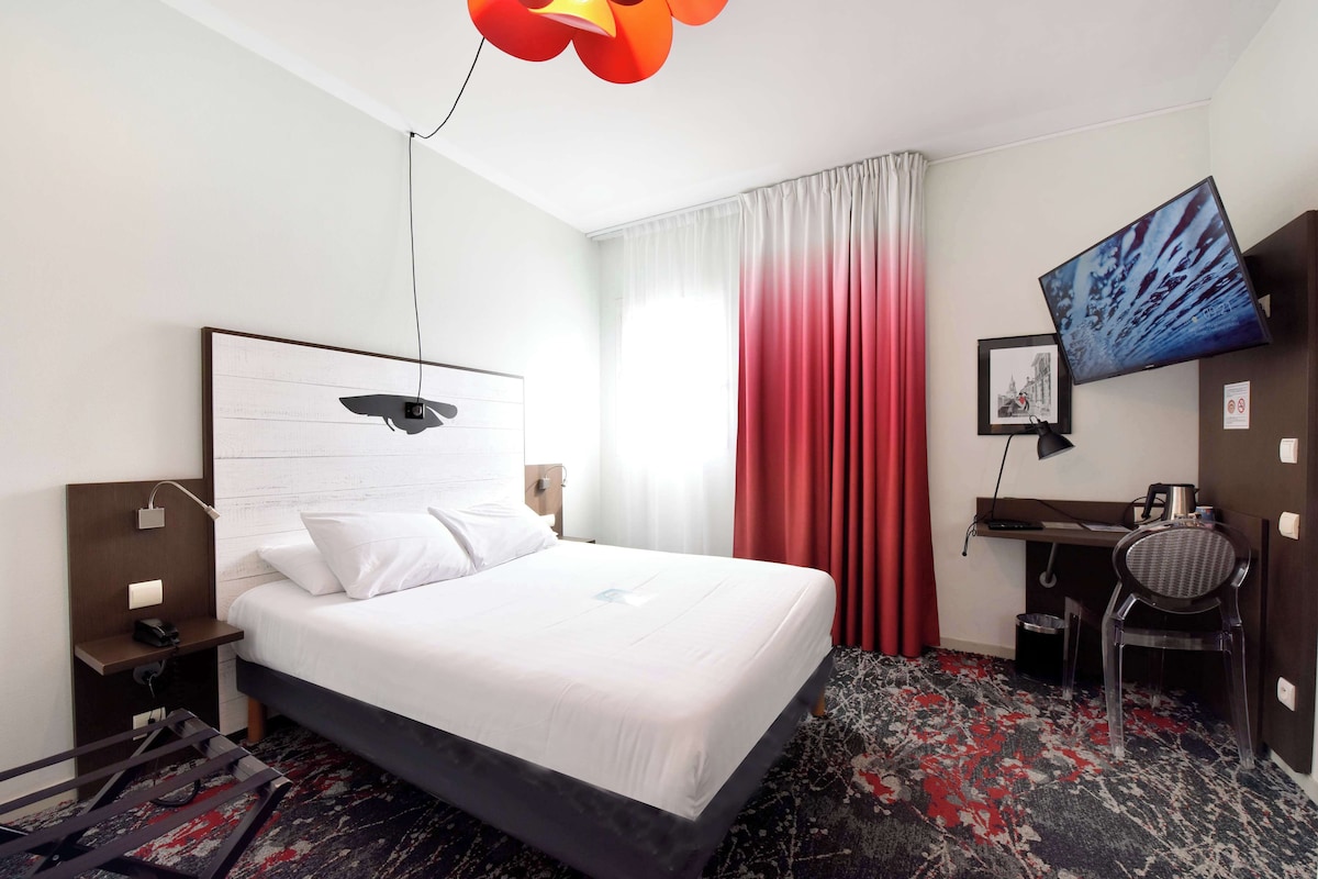 A hotel room with a bed, desk, chair, TV, and a red curtain.