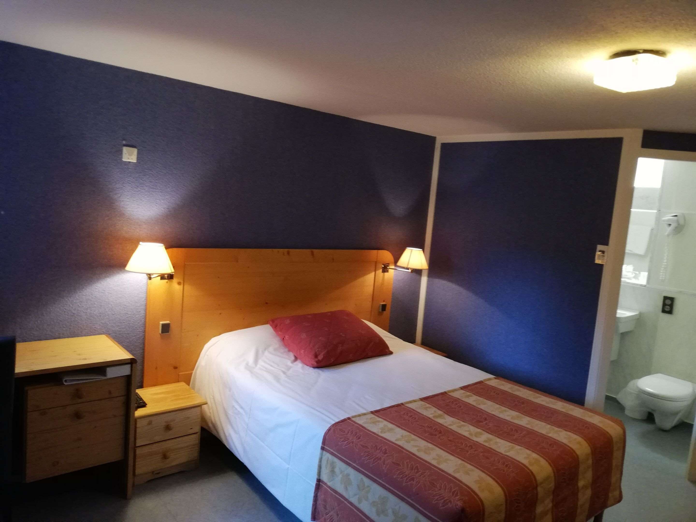 Een hotelkamer met een bed, bedlampen, een houten nachtkastje, een bureau en een badkamer zichtbaar door een geopende deur.