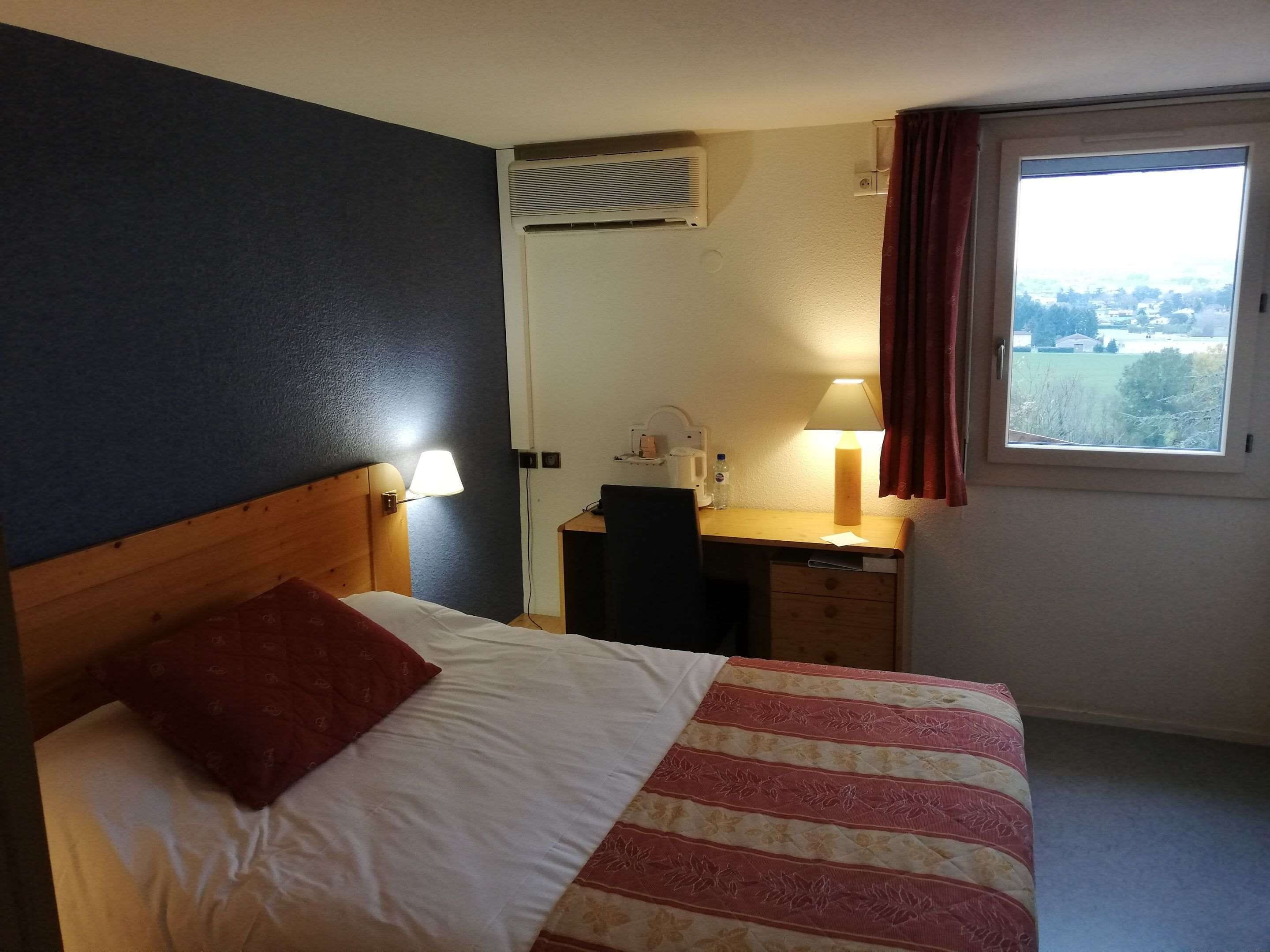 Habitación de hotel con cama, escritorio, silla, lámpara, ventana con cortinas y aire acondicionado.