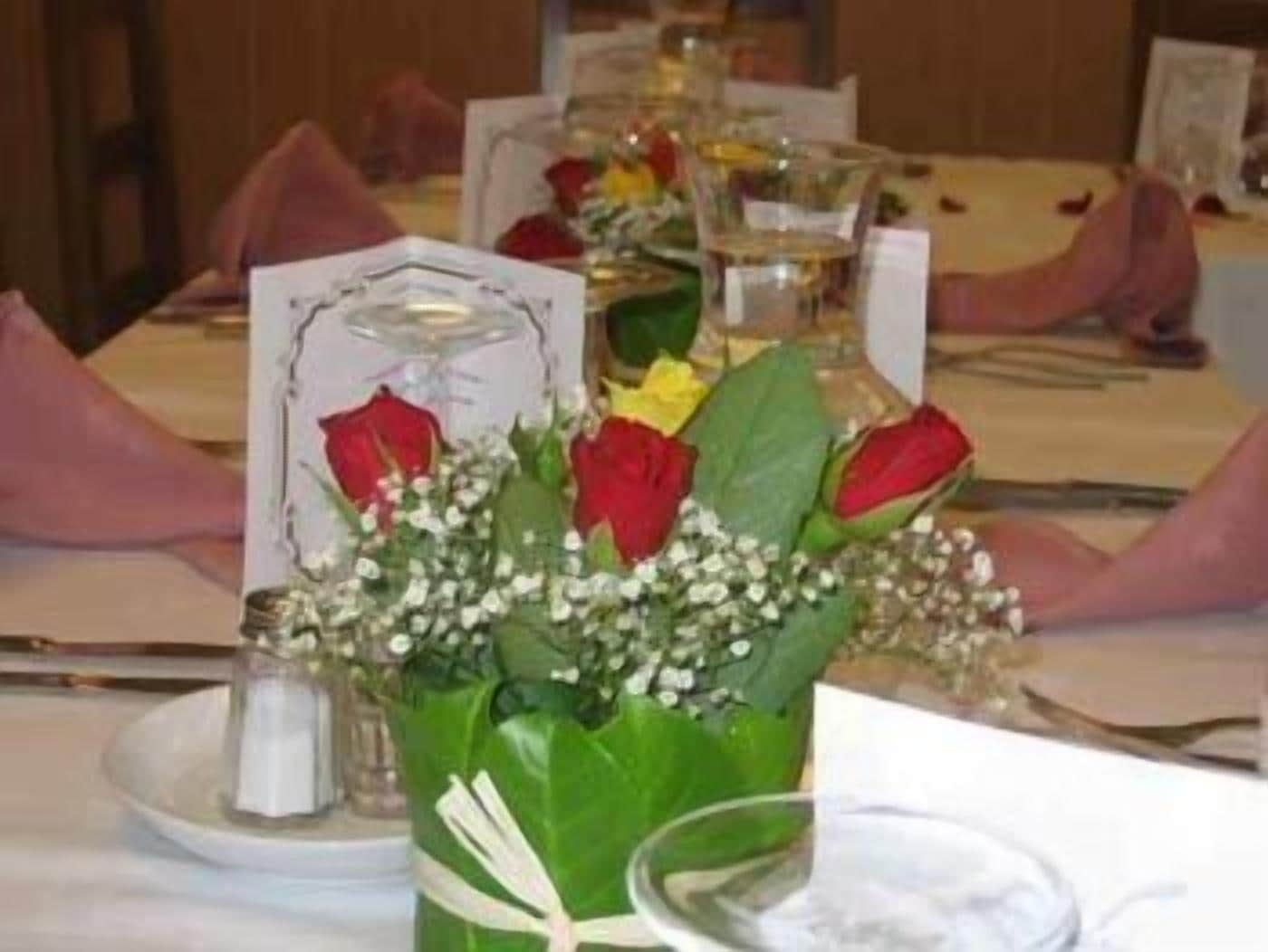 Ein gedeckter Tisch mit einer Mittelstückanordnung aus roten und gelben Rosen, weißen Blüten und einer Glasvase.