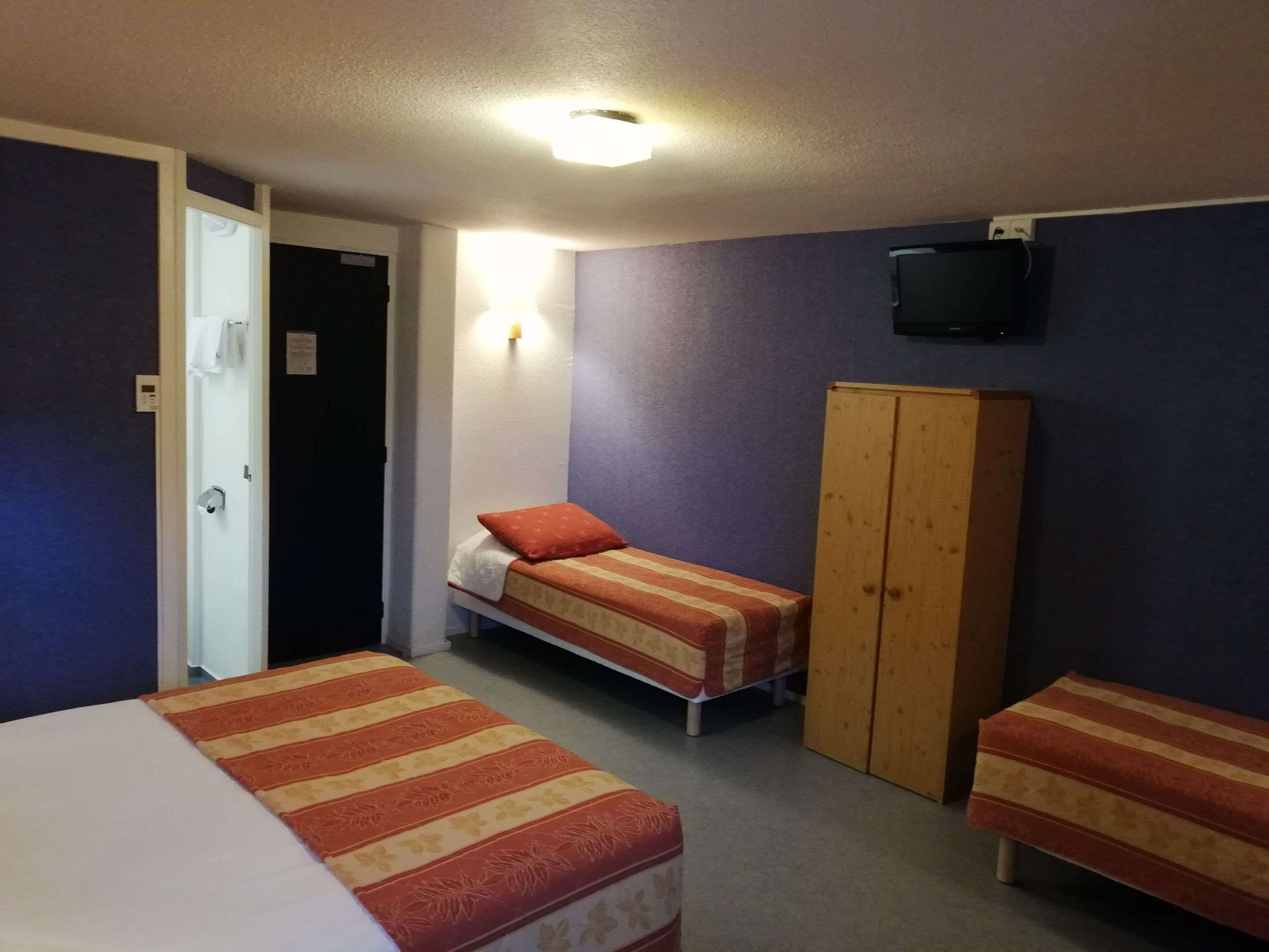 A room with two single beds, a wooden wardrobe, a television on the wall, and a door leading to another room.