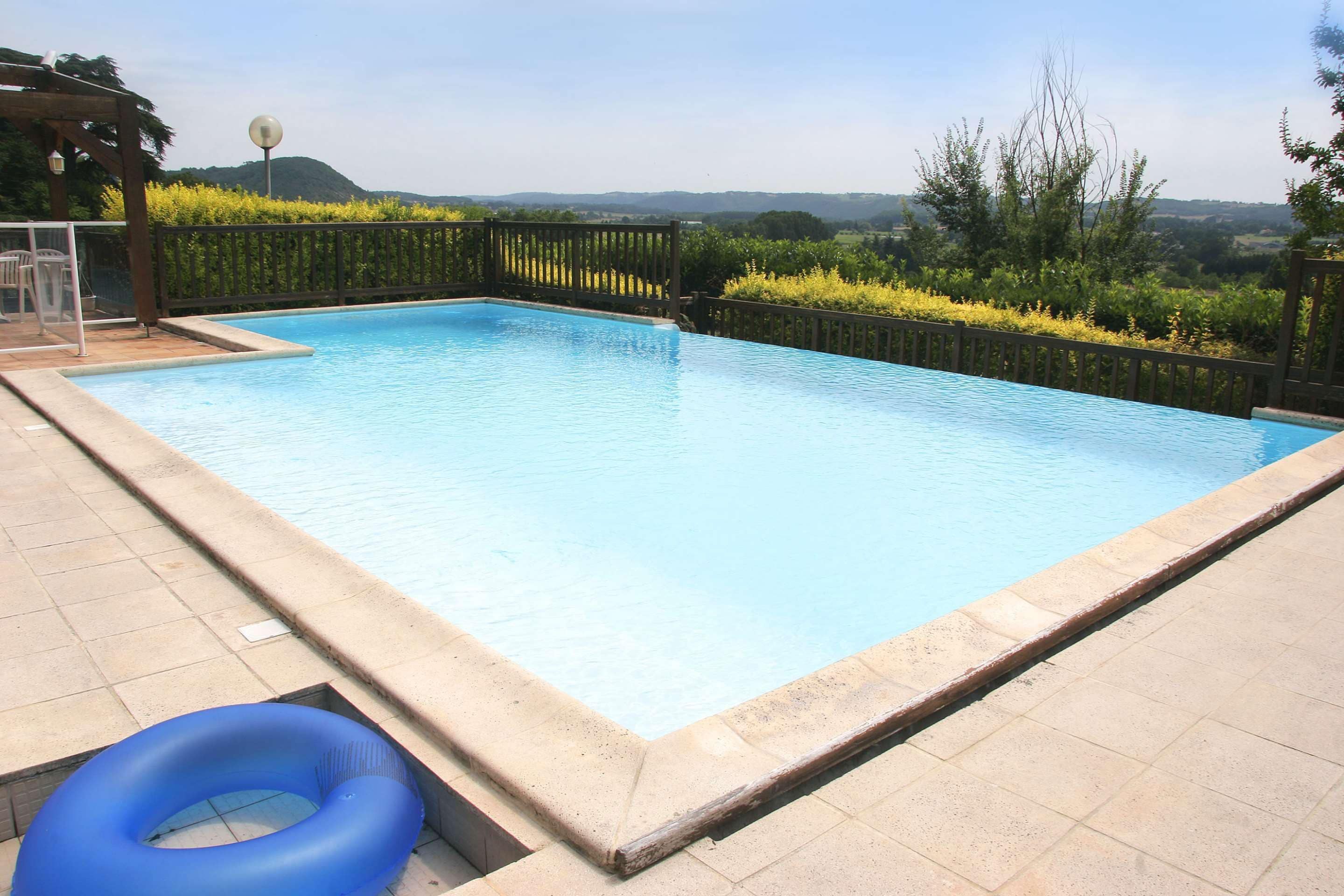 A rectangular swimming pool with a tiled deck, surrounded by a wooden fence and greenery.