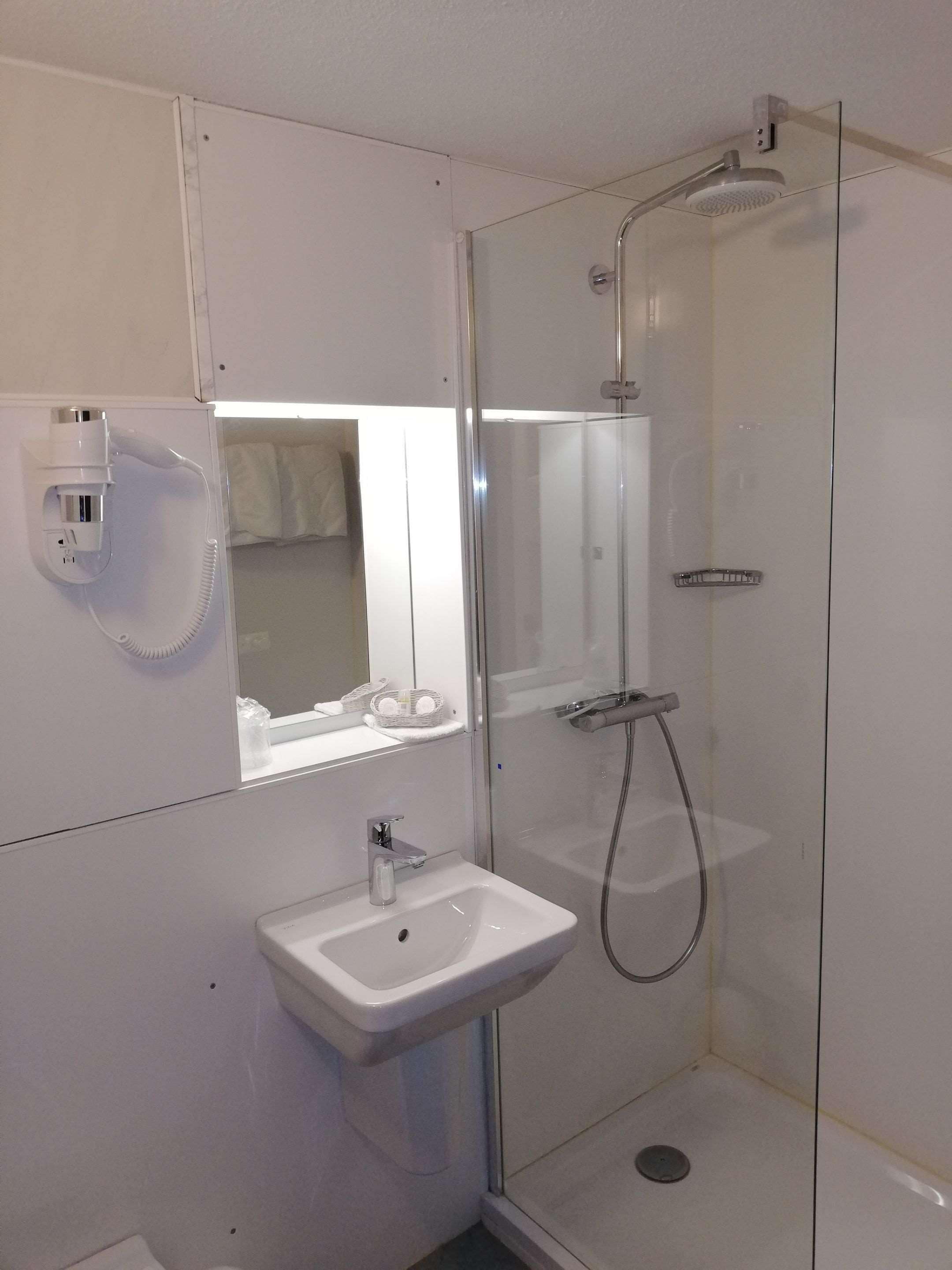 Un baño moderno con ducha envuelta en vidrio, lavamanos blanco y un secador de pelo fijado en la pared.