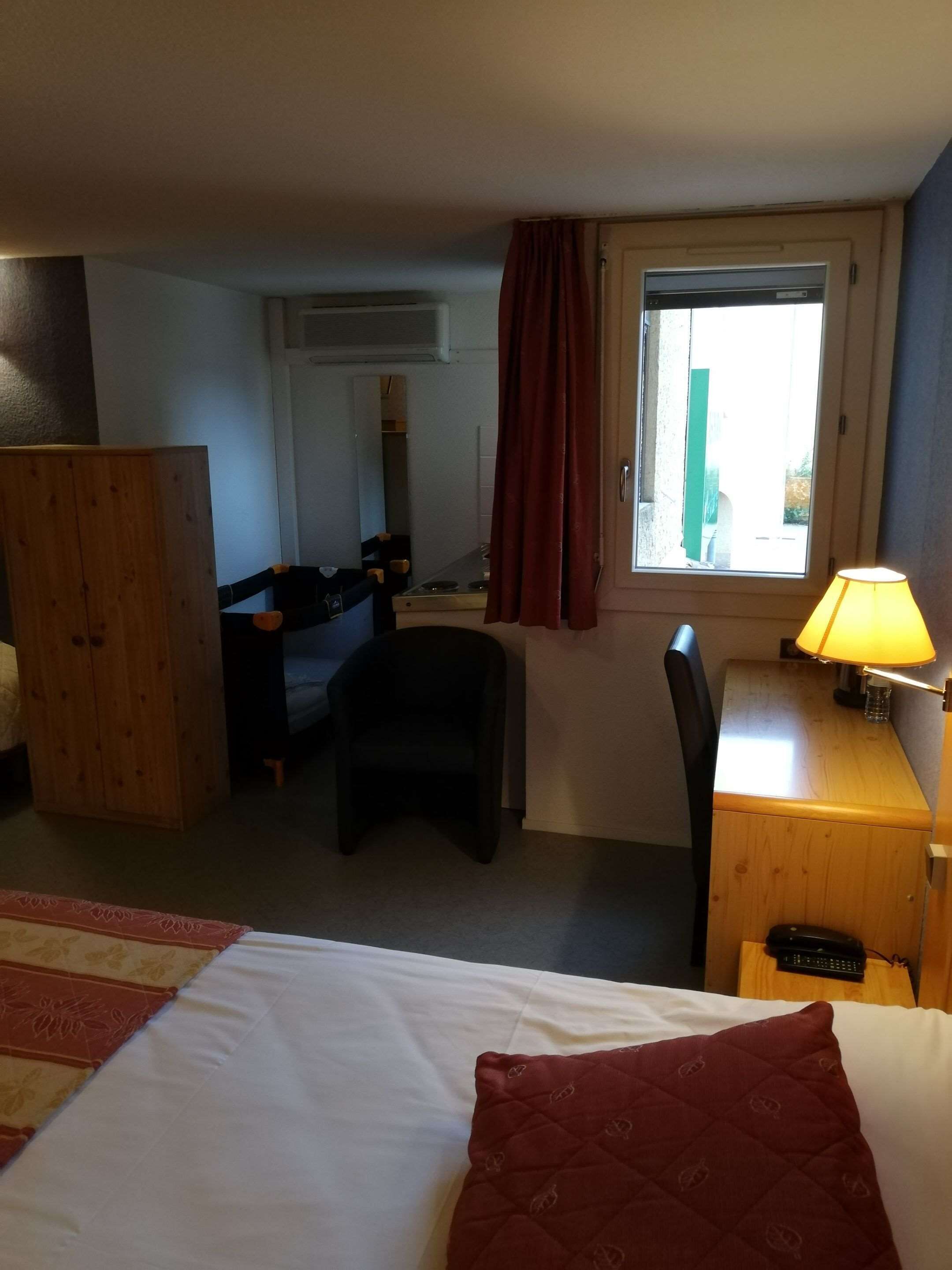 Una habitación de hotel con una cama, un escritorio con lámpara, una silla, un armario y una ventana con cortinas.