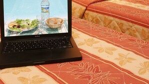 Um laptop sobre a cama, que tem cabeceira de madeira, uma luminária e um edredom estampado.