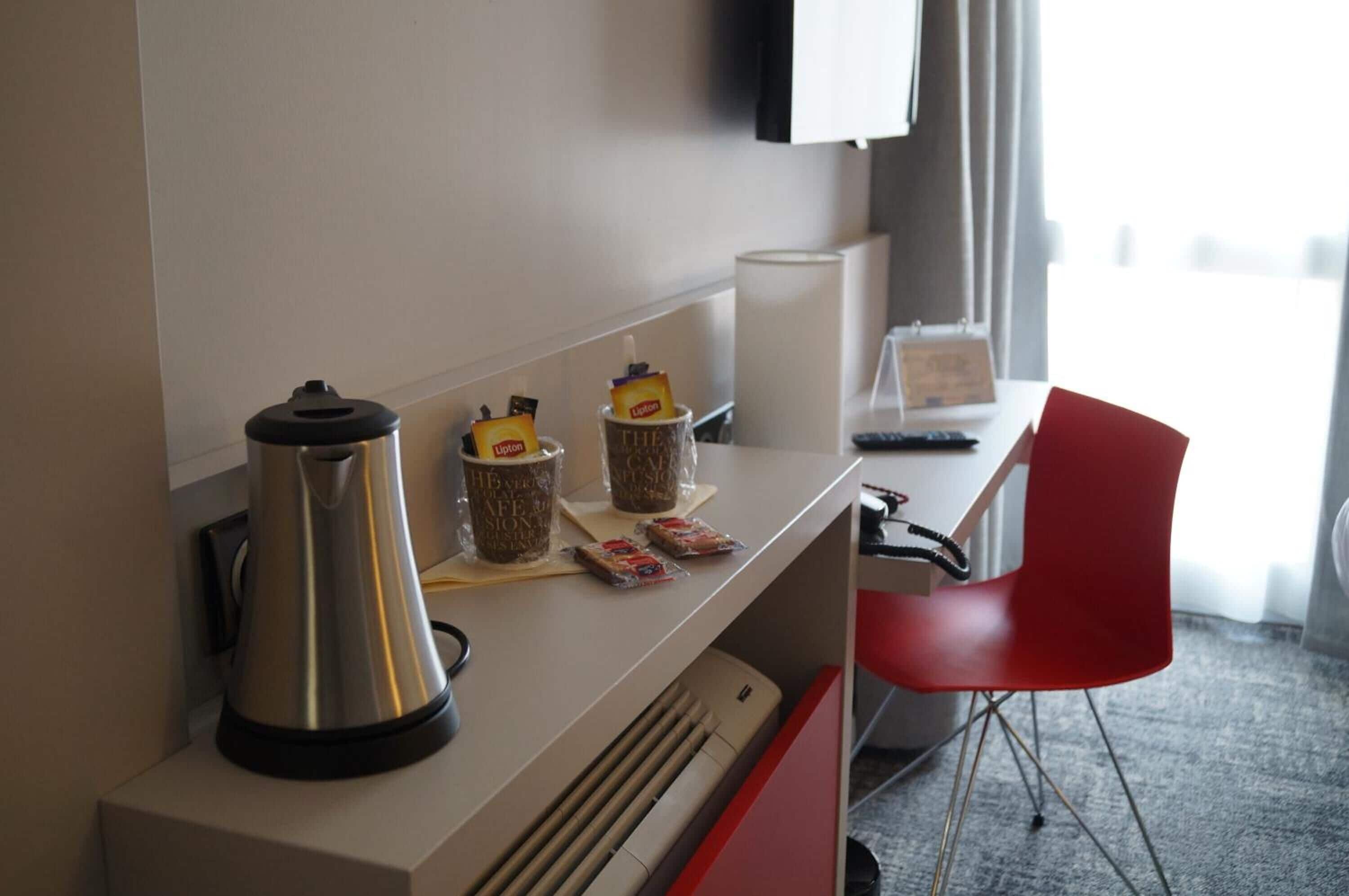 Habitación de hotel con kitchenette equipada con cafetera, tostadora y un refrigerador pequeño. Se observa una silla roja y un televisor de pantalla plana montado en la pared.