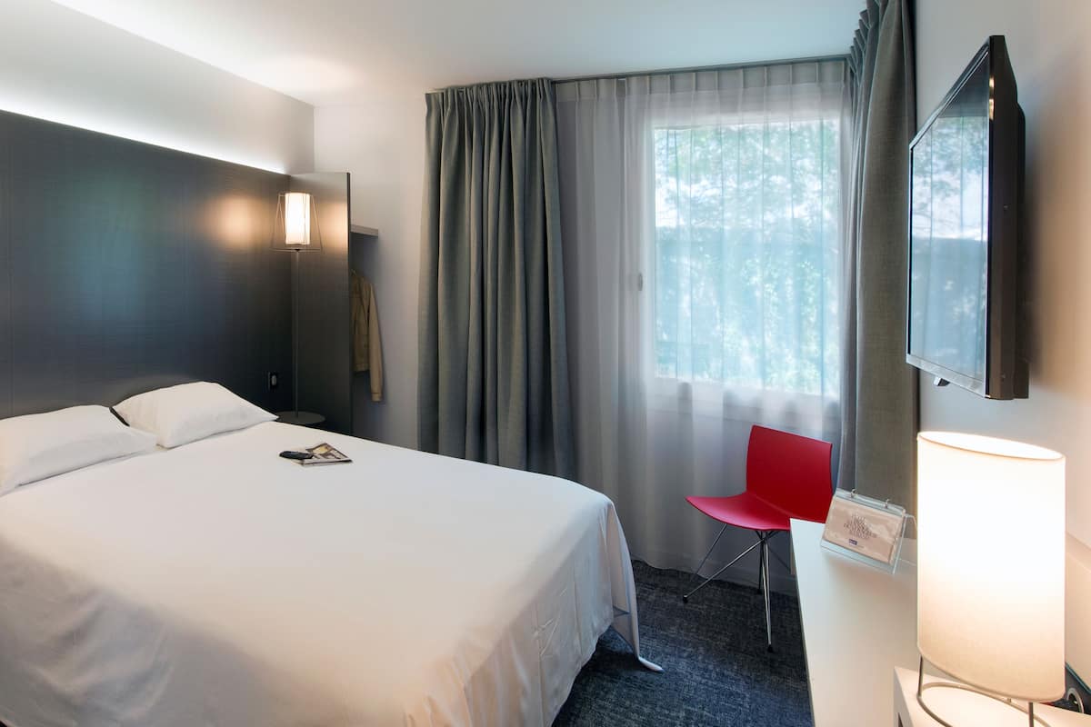 Una habitación de hotel con una cama, una silla roja, un escritorio con lámpara y un ventanal con cortinas.