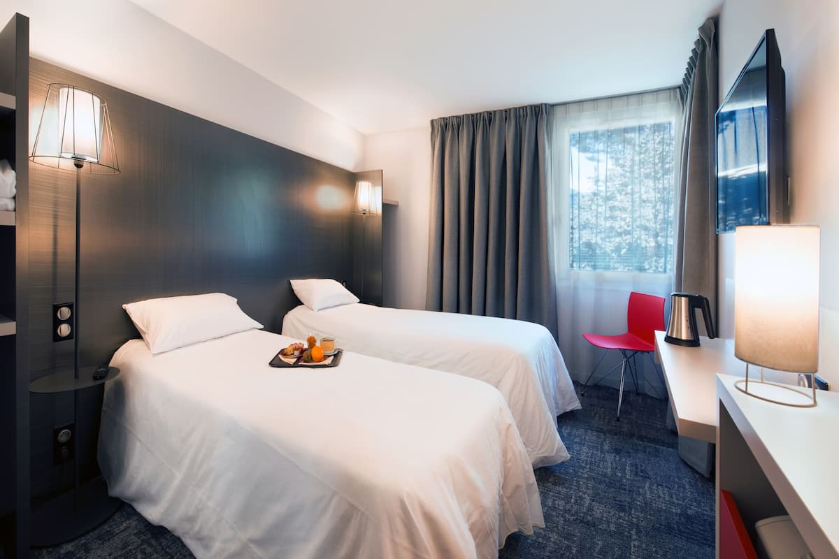 Habitación de hotel con dos camas, un escritorio, una silla, una lámpara, un televisor y una ventana con cortinas.