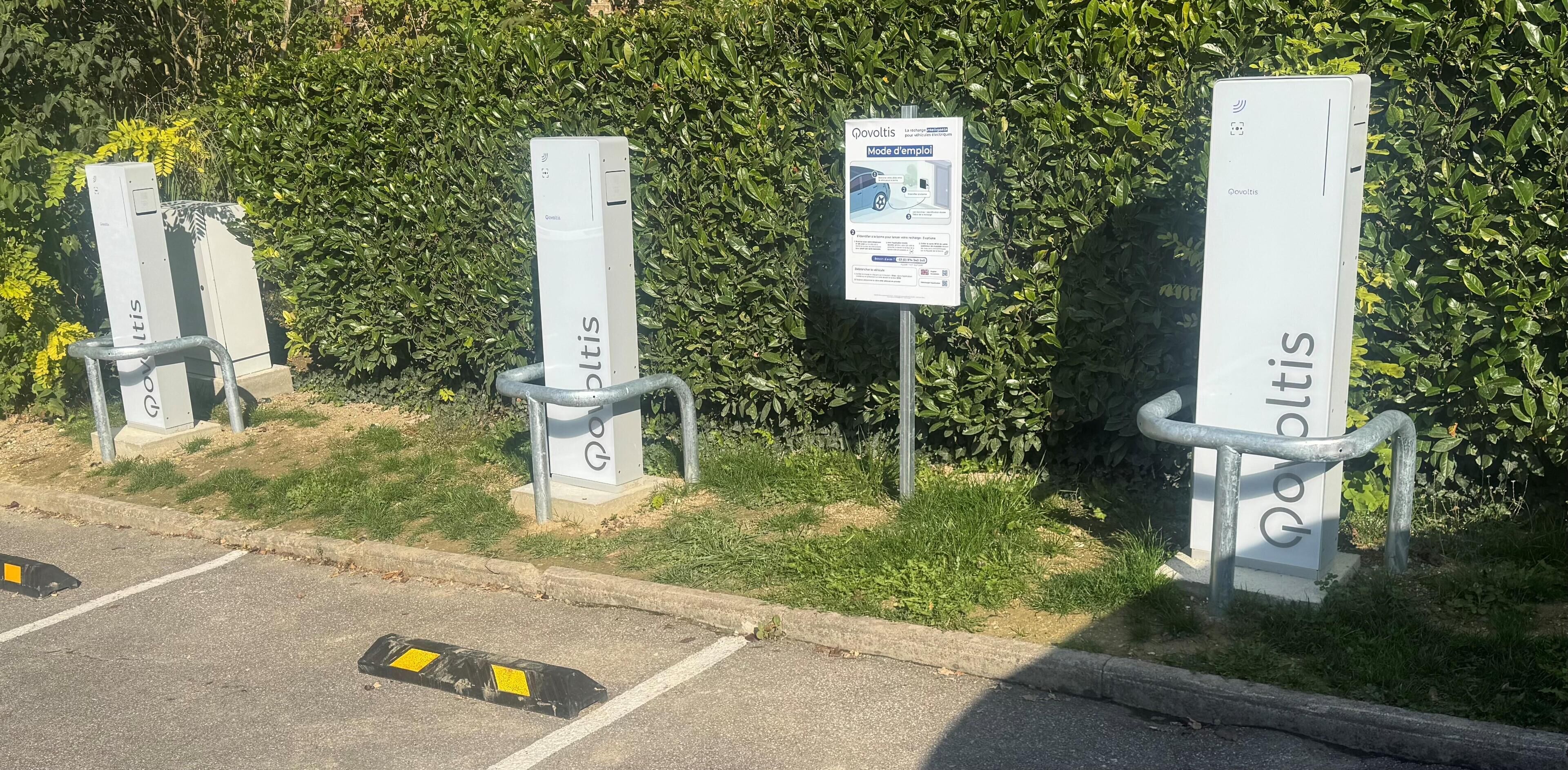 Electric vehicle charging stations with metal railings, labeled 'Povoltis,' against a backdrop of green foliage.