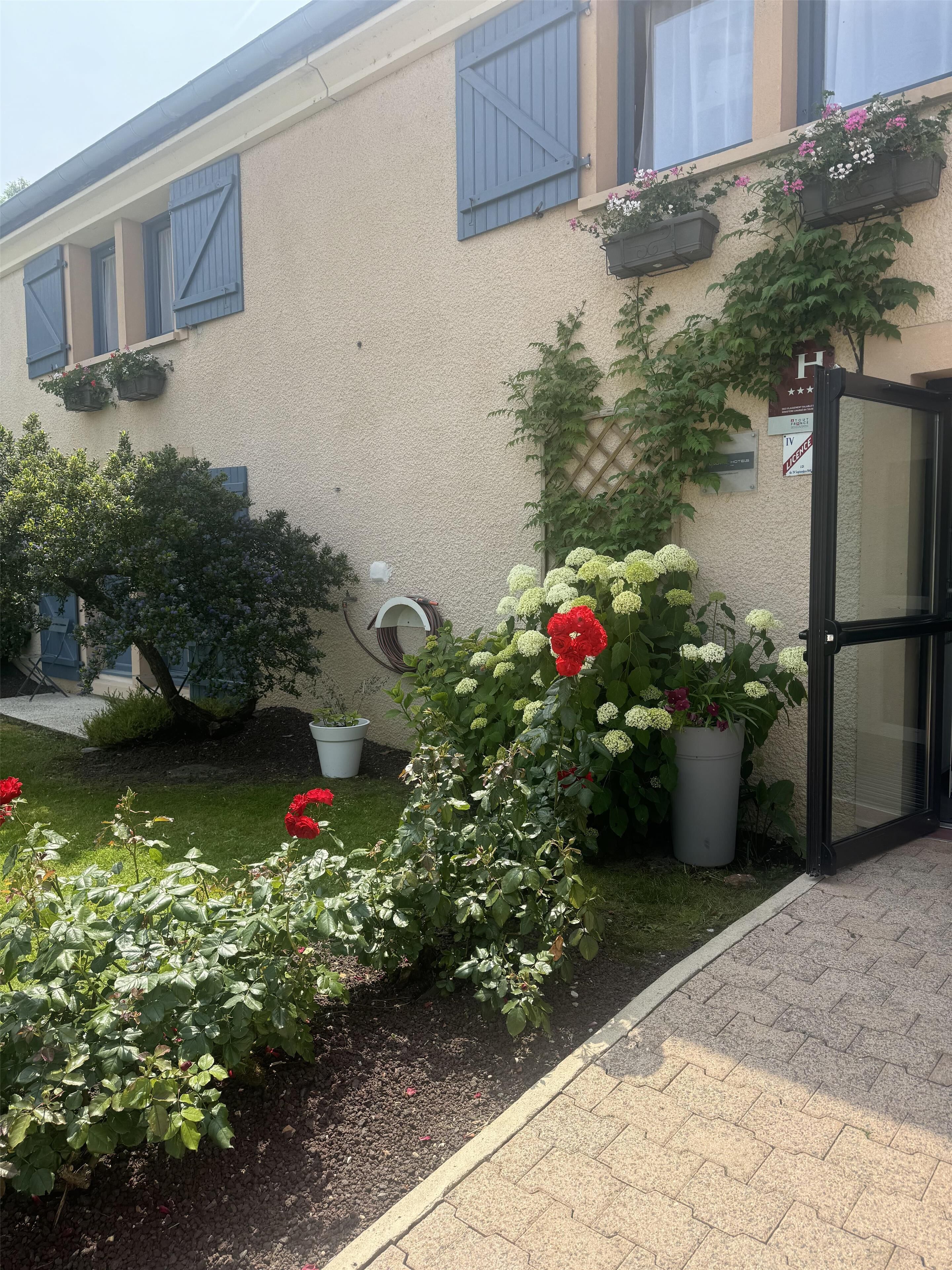 Un bâtiment à la façade beige, avec des volets bleus et des jardinières sur les rebords des fenêtres. On y trouve une zone pavée agrémentée de plantes et de fleurs en pot.