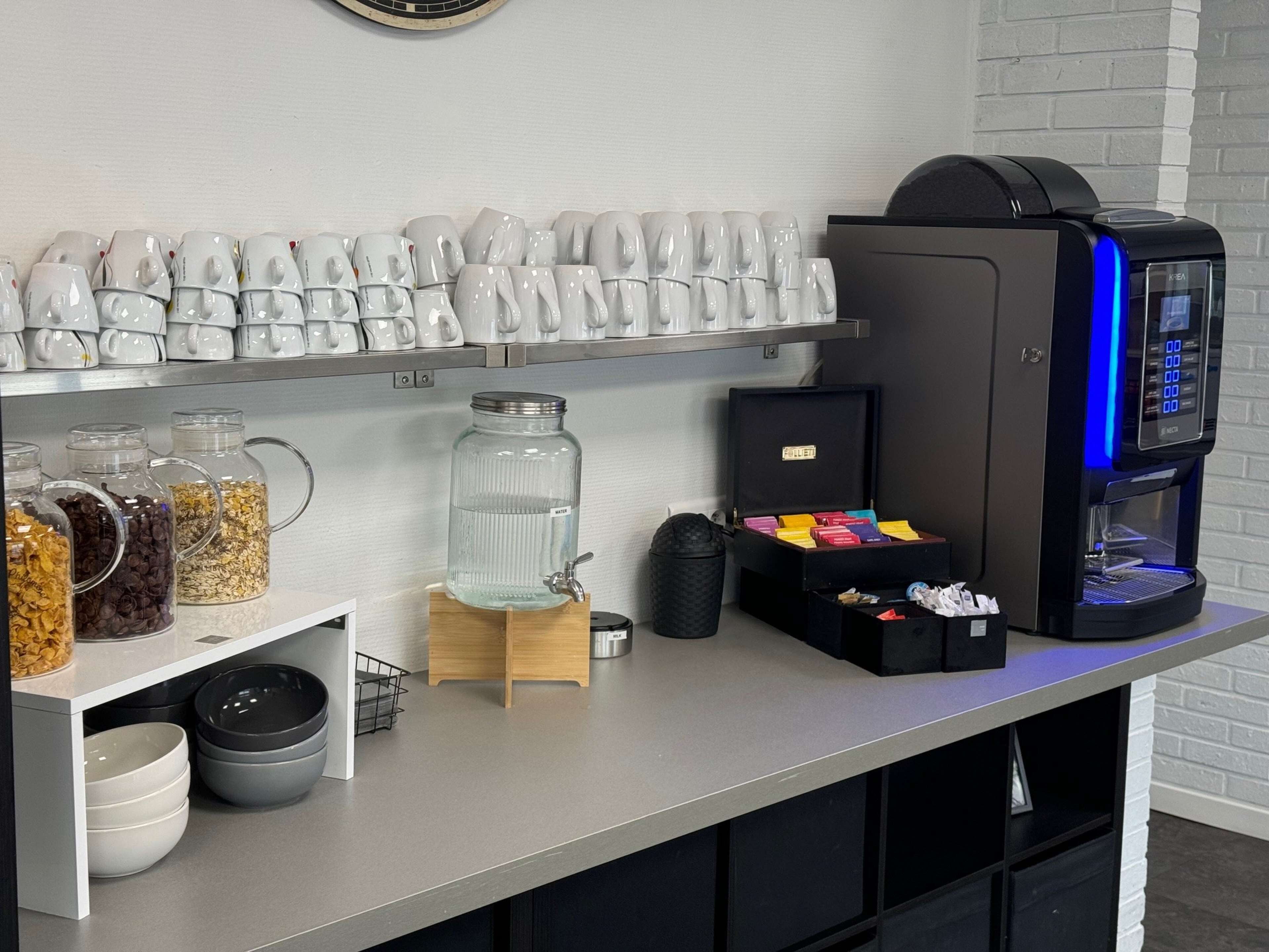 Una estación de café con una máquina de café, tazas y varios recipientes de comida.