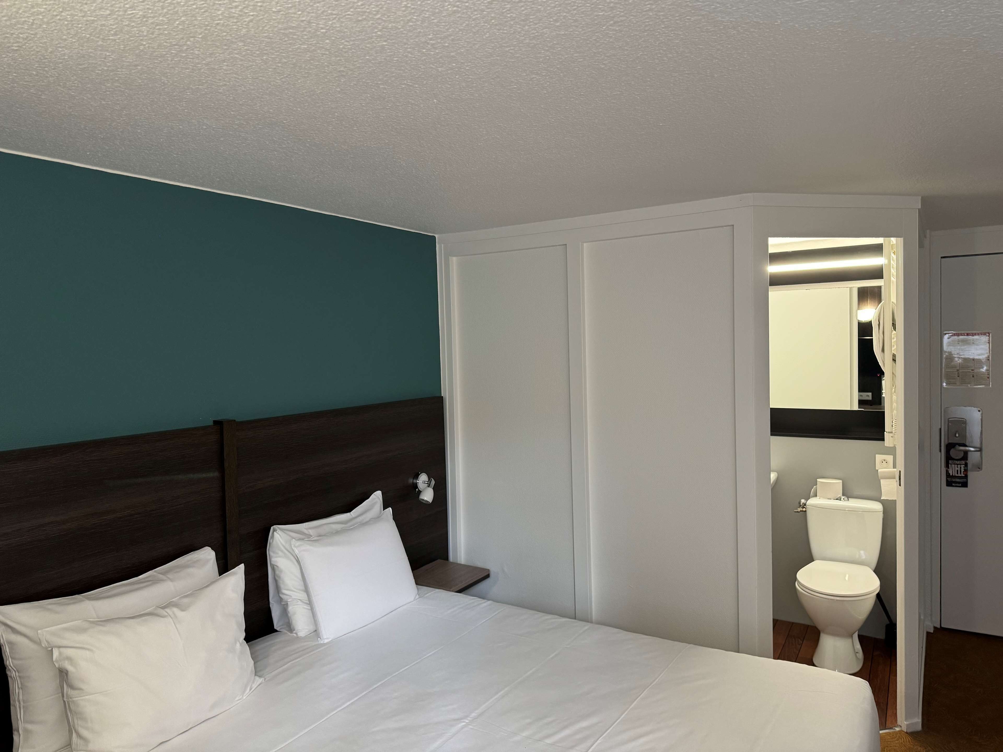 Quarto de hotel com cama, mesa de cabeceira, sanita branca e espelho.