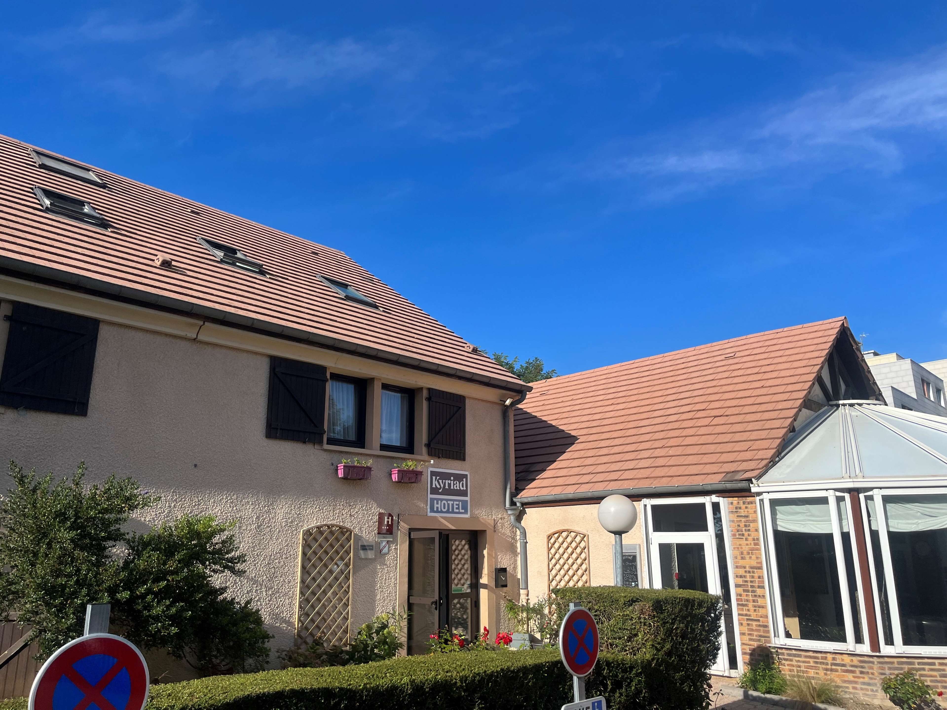 A hotel with a tiled roof, a sign indicating 'Kyriad Hotel', and a no parking sign.