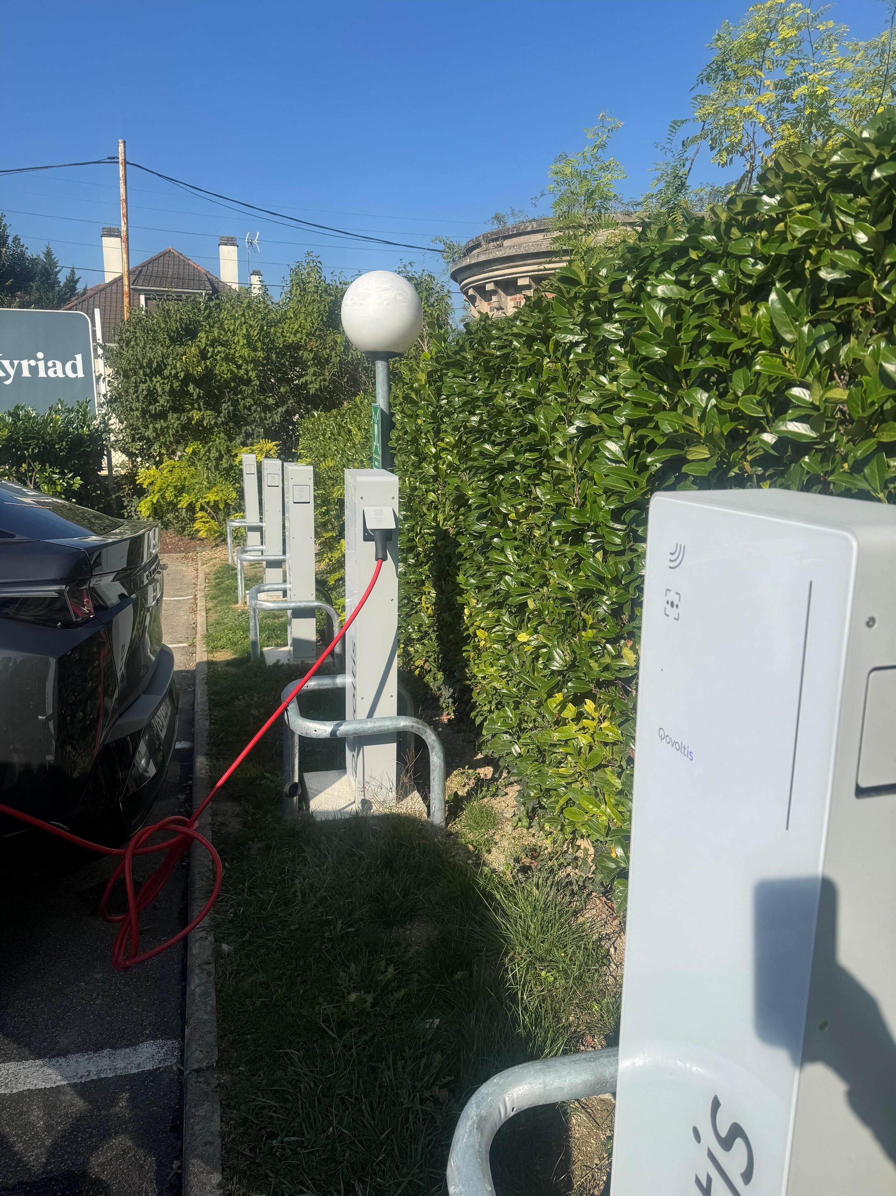 Un espace de stationnement équipé de bornes de recharge pour véhicules électriques, une voiture et un bâtiment en arrière-plan.