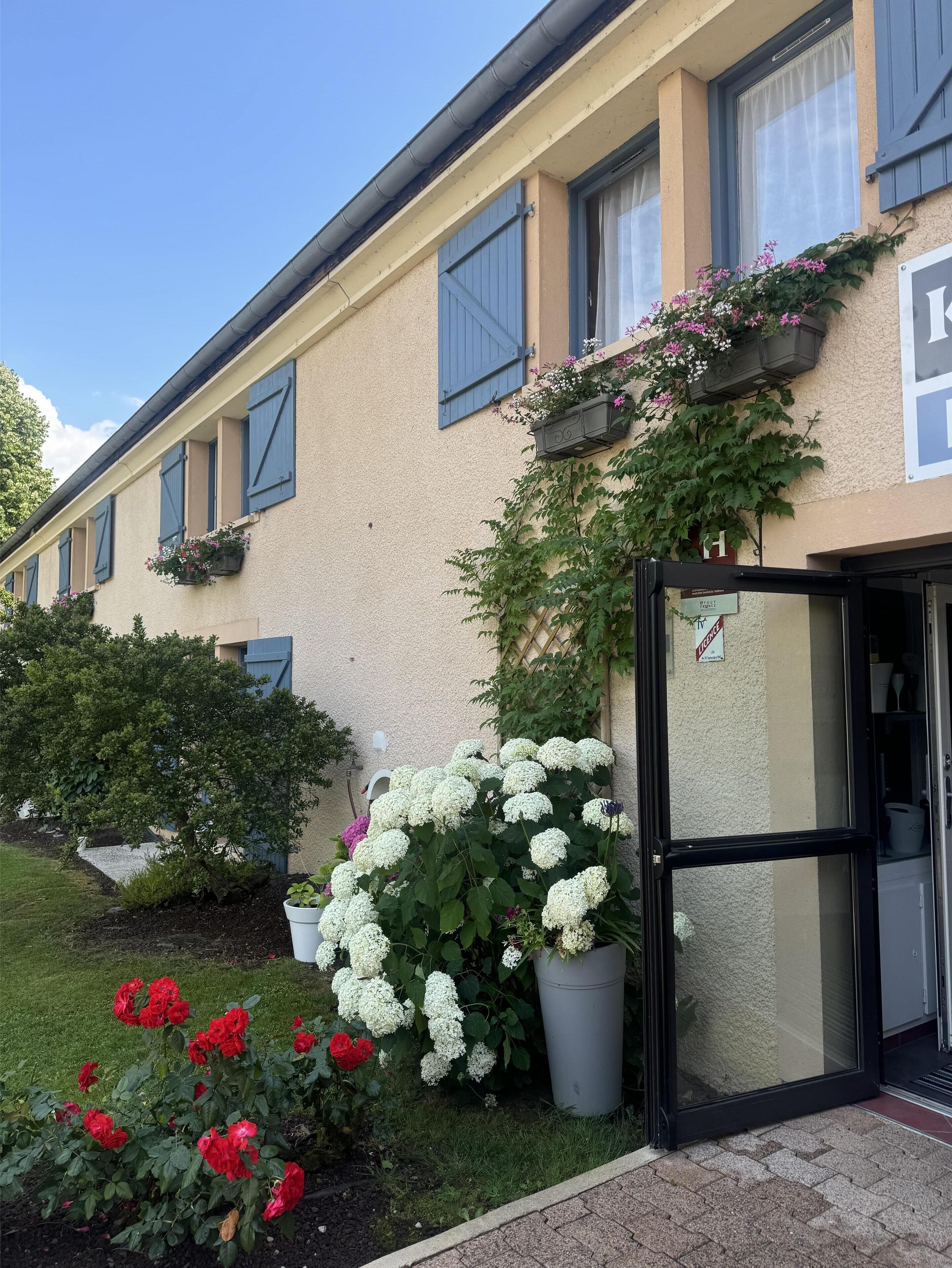 A building with a beige facade, blue shutters, and a garden with white and red flowers.