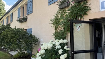 Un edificio con fachada color crema, contraventanas azules y un jardín con flores blancas y rojas.