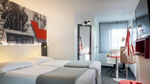 A hotel room with two beds, a desk, a chair, a mirror, and a red stool.