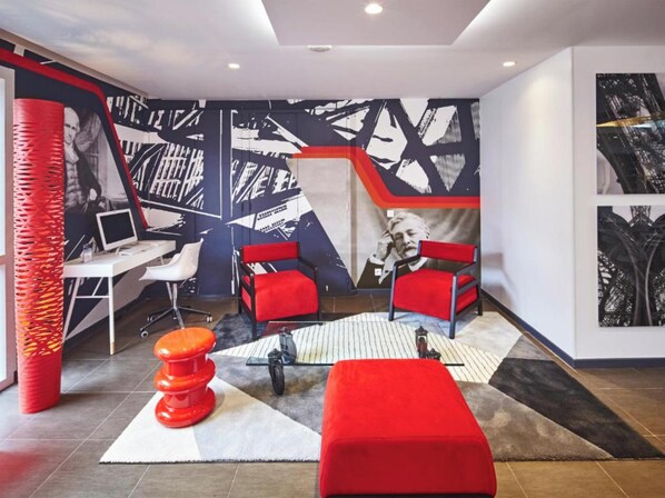 A modern room with a desk, two red chairs, a red ottoman, and a large abstract mural on the wall. - Hôtel Gustave Colmar (Colmar)