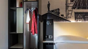 A hotel room with a bed, a coat rack with a red plaid shirt, a shelf with books, and a wall with a black and white photo.
